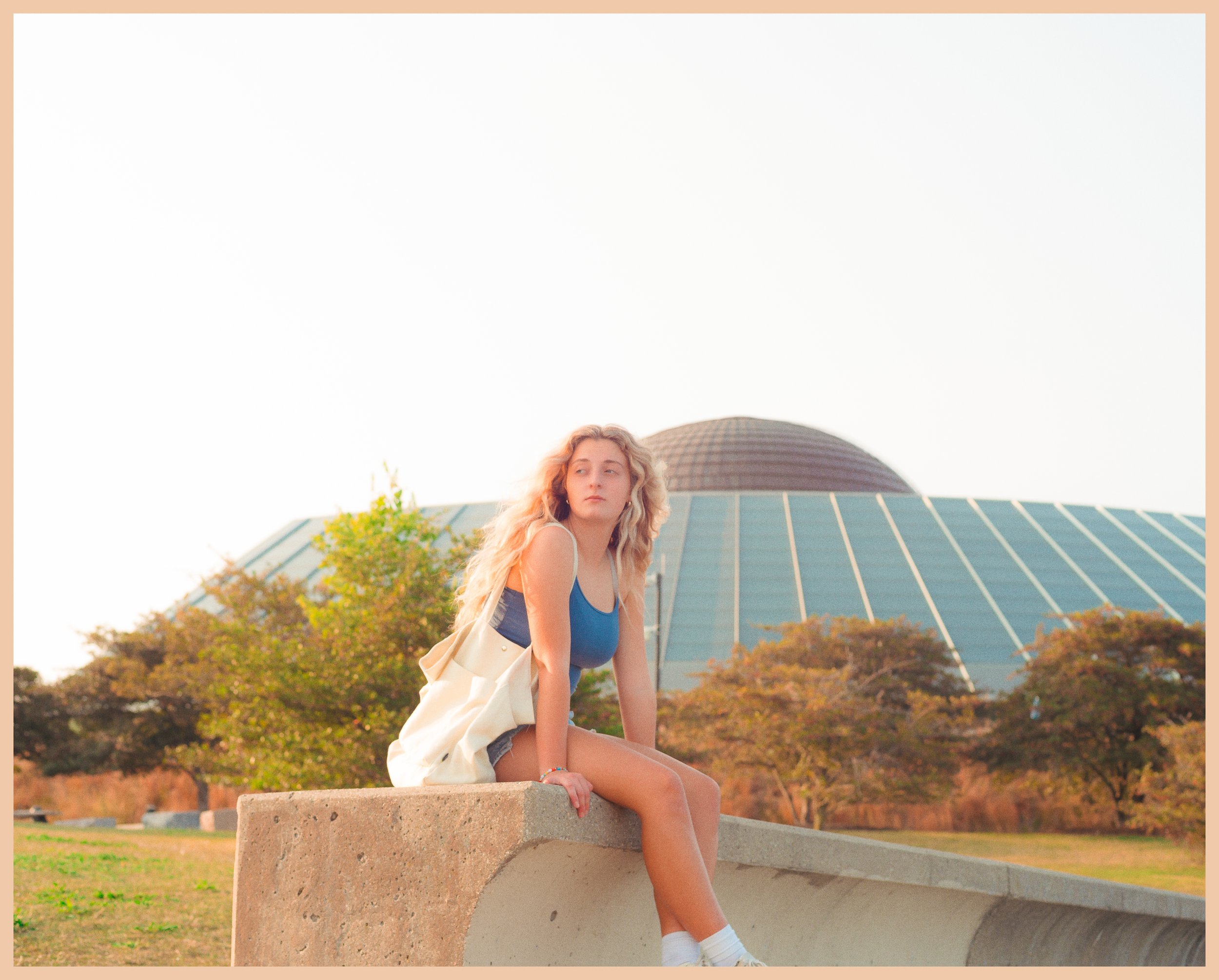 A young woman with long, wavy blonde hair sits on a concrete ledge outdoors during sunset, with a modern, dome-shaped building in the background and trees with fall foliage around her.