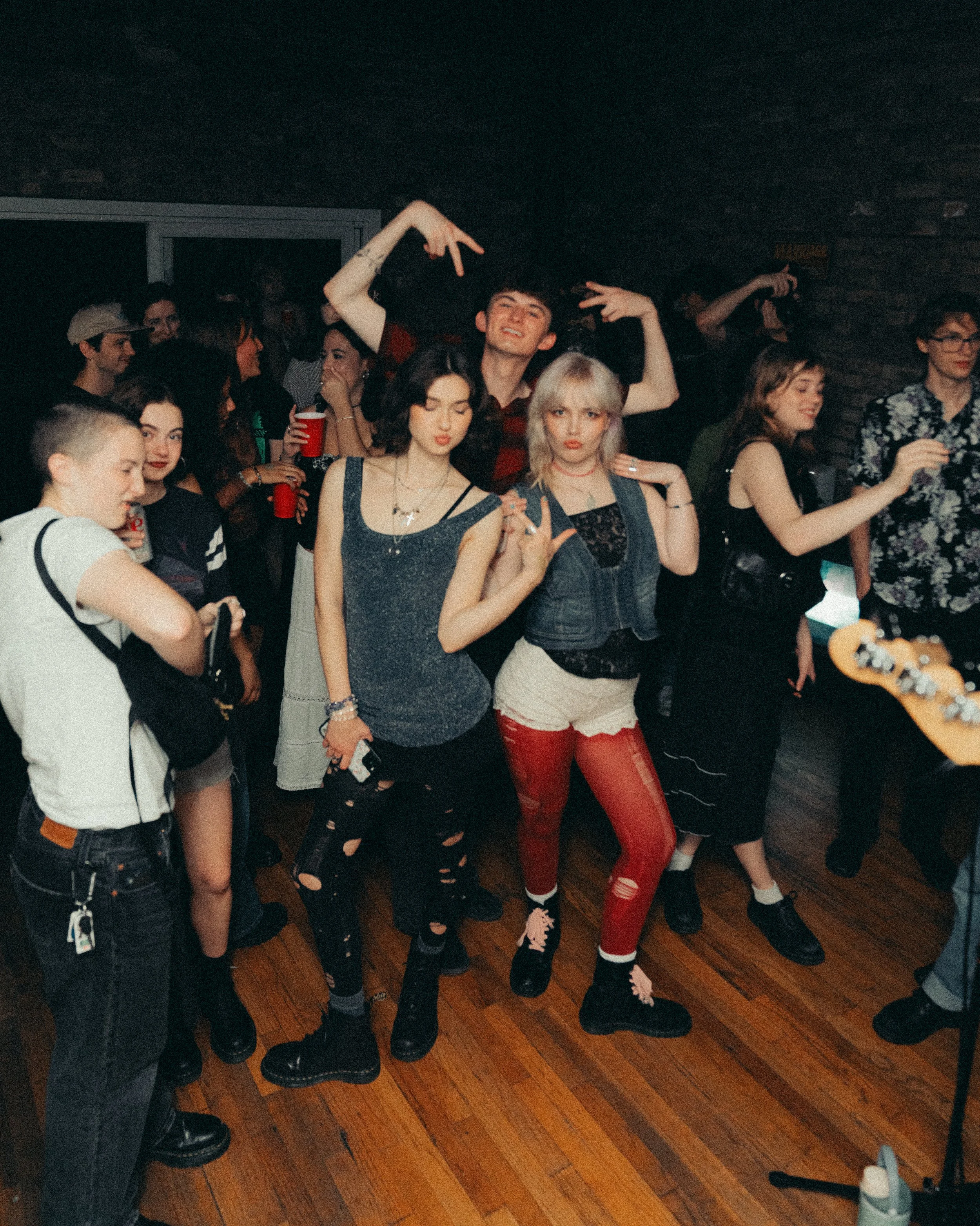 Group of young people at a party or concert, dancing and having fun indoors with a dark ambiance and wooden floor.
