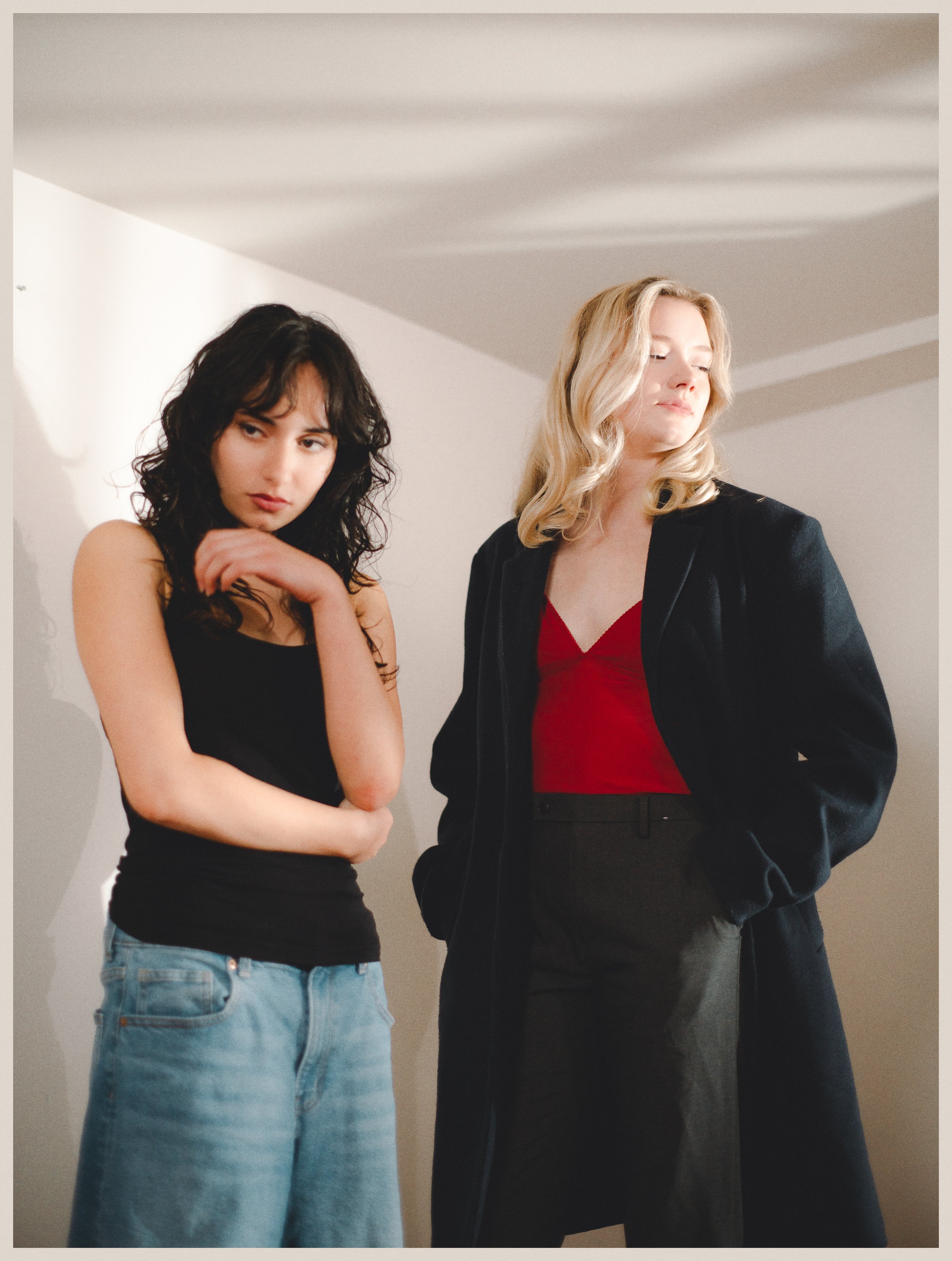 Two women standing indoors against a neutral background, with one woman having dark curly hair and wearing a black sleeveless top and jeans, and the other woman having blonde wavy hair and wearing a red top with a black blazer and black pants.
