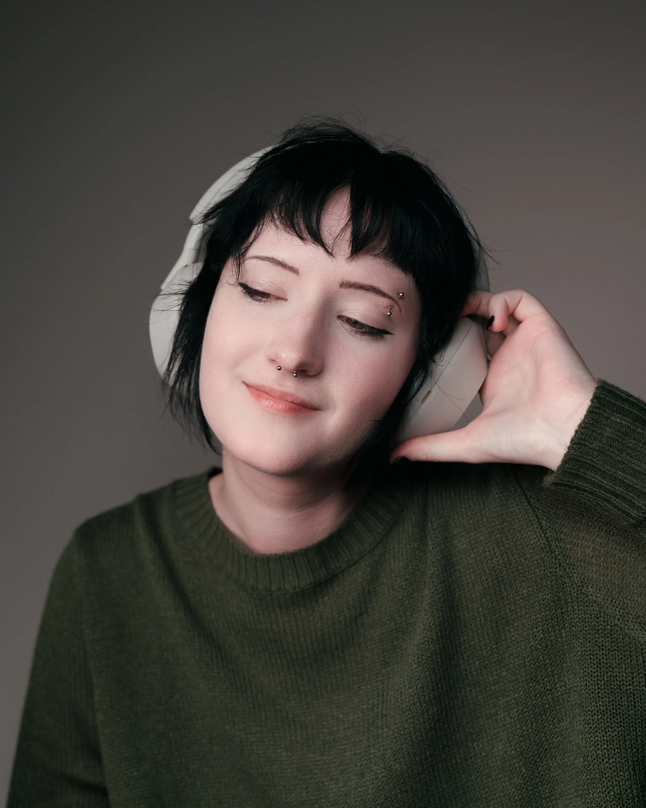 Young woman with black hair, piercings, and closed eyes enjoying music through white headphones, wearing a dark green sweater against a plain background.