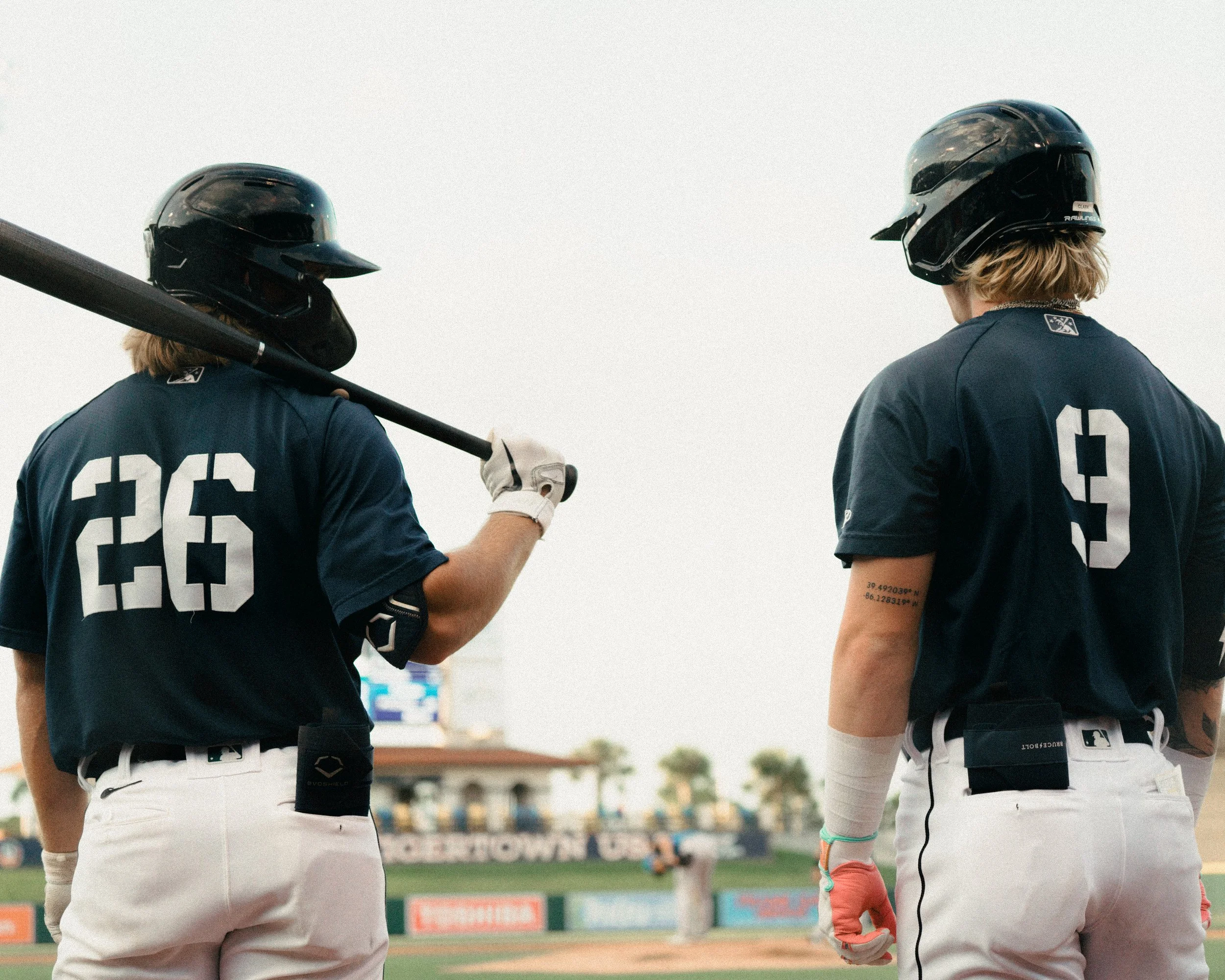 Two baseball players in navy blue jerseys and white pants standing on the field, one holding a bat over his shoulder, with a baseball stadium in the background.