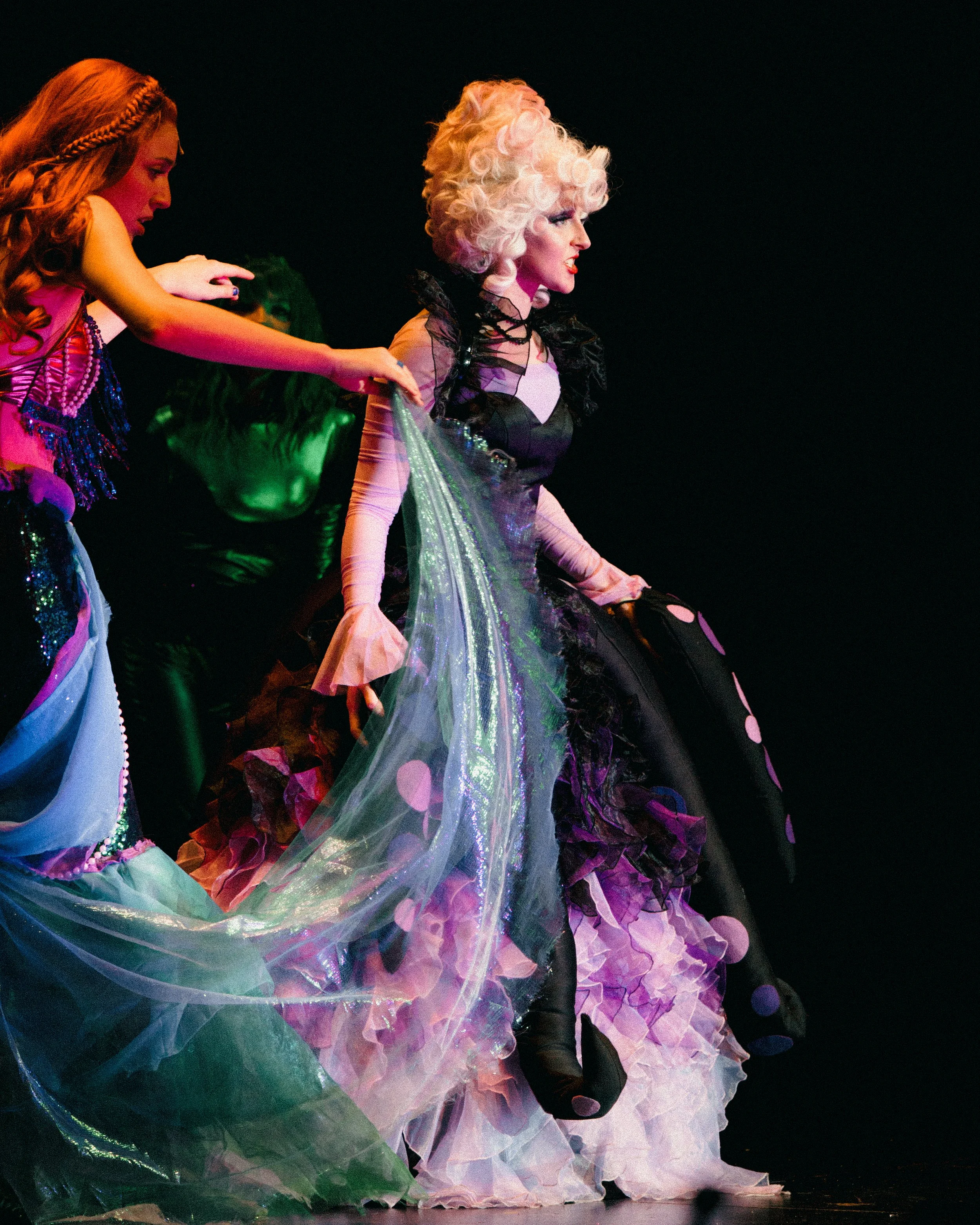 A stage performance featuring performers in elaborate, colorful costumes, with one person in a black and pastel gown with striped shoes and curly white hair.