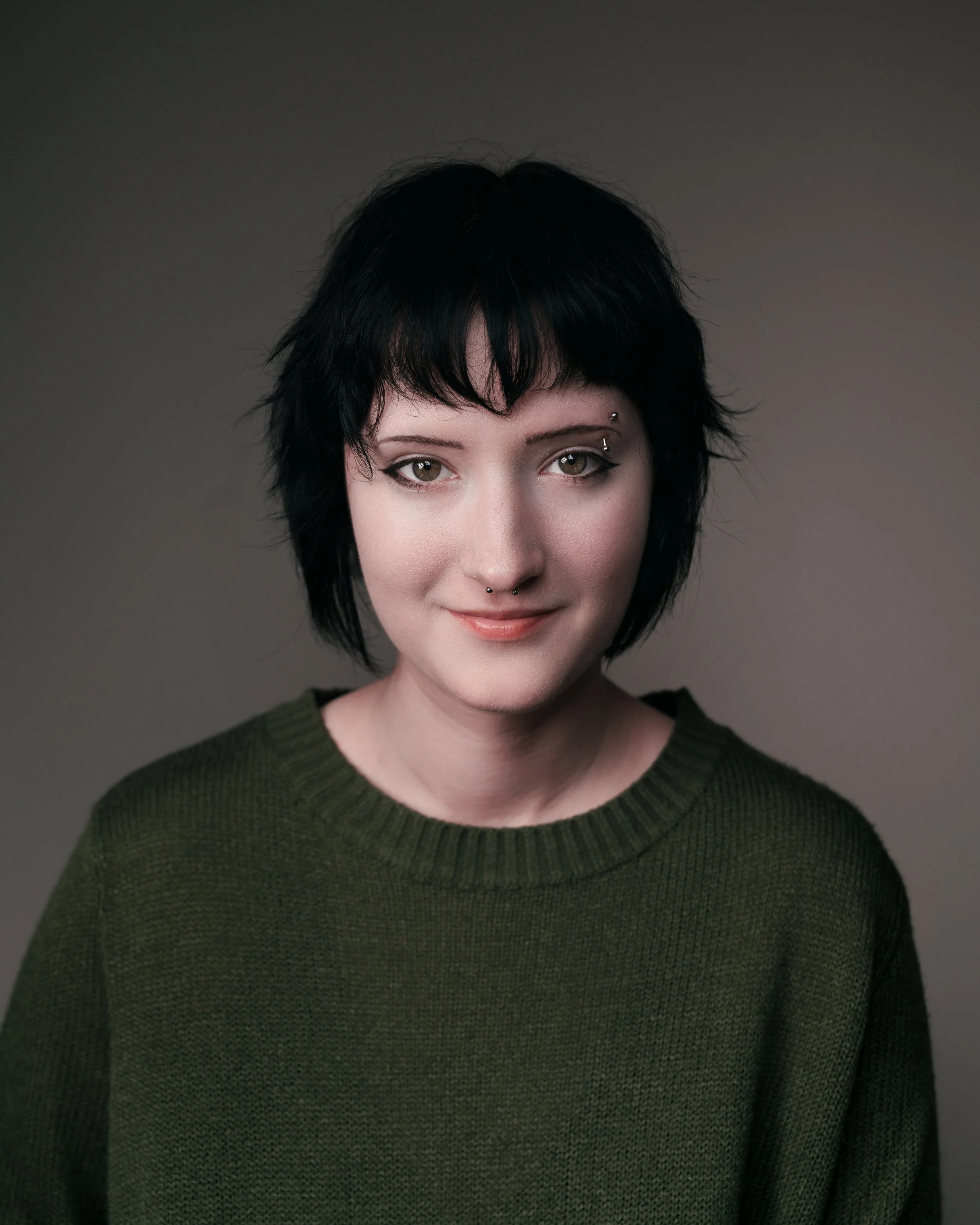 A young woman with short black hair, light eye makeup, and facial piercings, wearing a dark green sweater, smiling at the camera against a gray background.