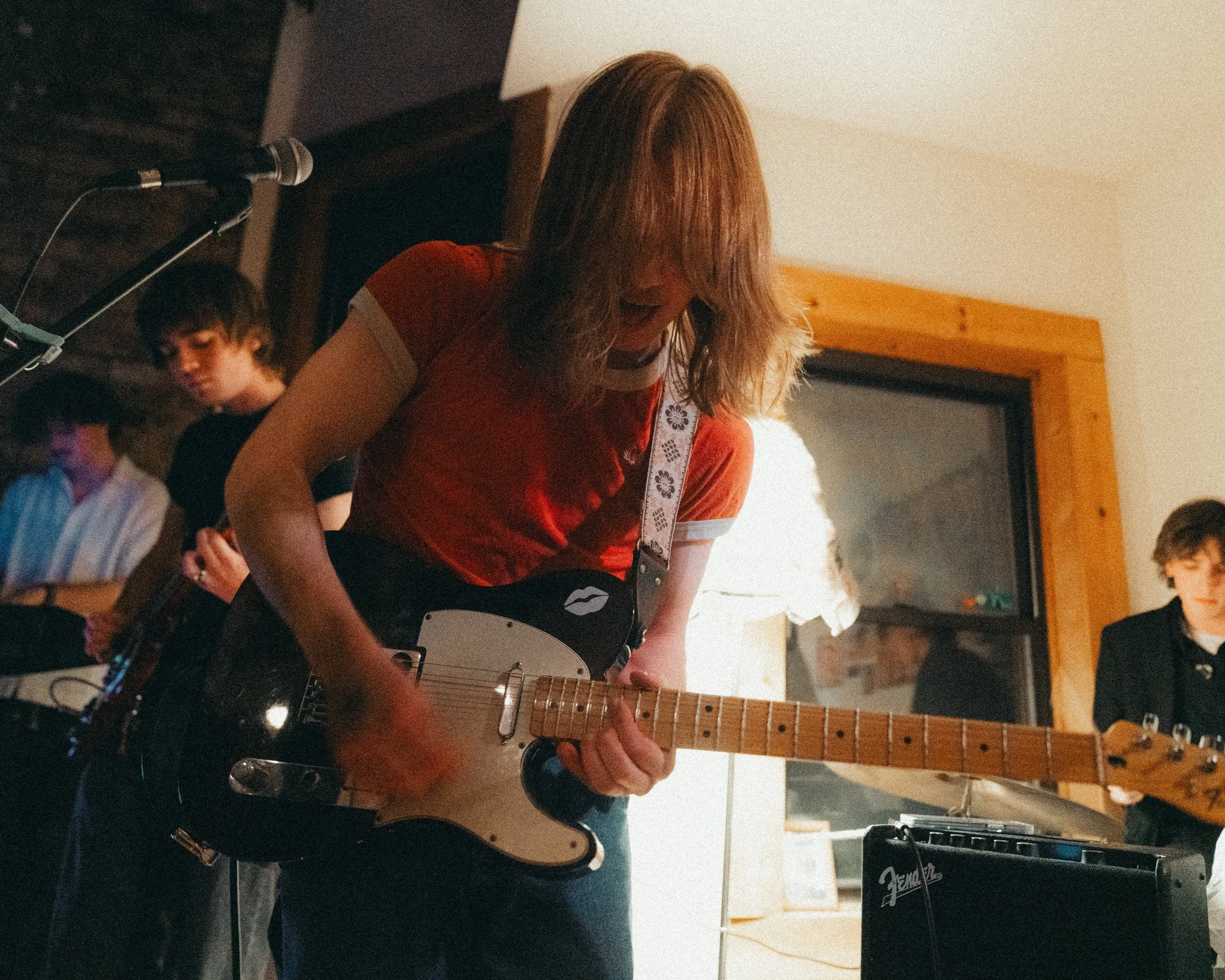 A woman playing an electric guitar during a band performance indoors, with two other band members visible in the background.