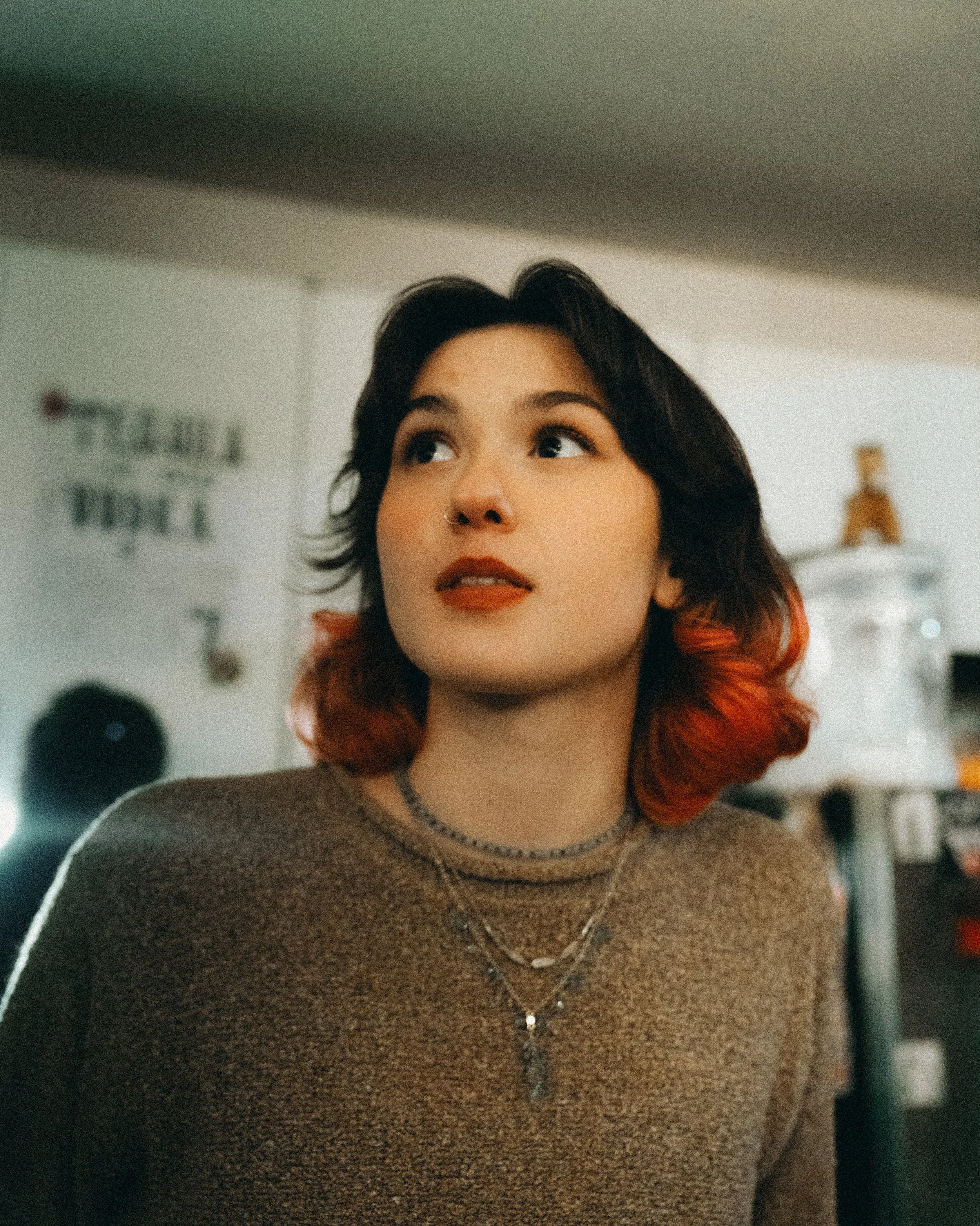 A young woman with shoulder-length dark hair with red tips, wearing necklaces and a brown sweater, looking upward in an indoor setting.
