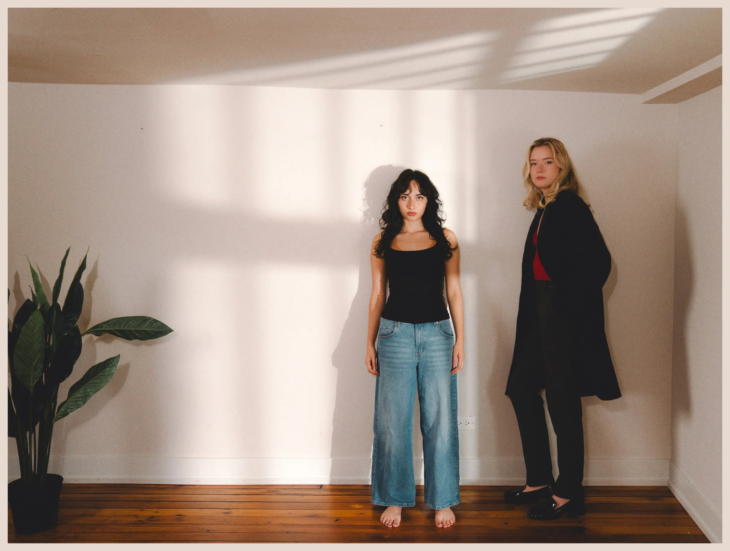 Two women stand barefoot against a plain white wall, with light casting shadows on the ceiling. One woman with dark curly hair wears a black tank top and blue jeans. The other woman with blonde hair wears a black blazer, black pants, and a red top. There is a potted houseplant on the left side of the image.