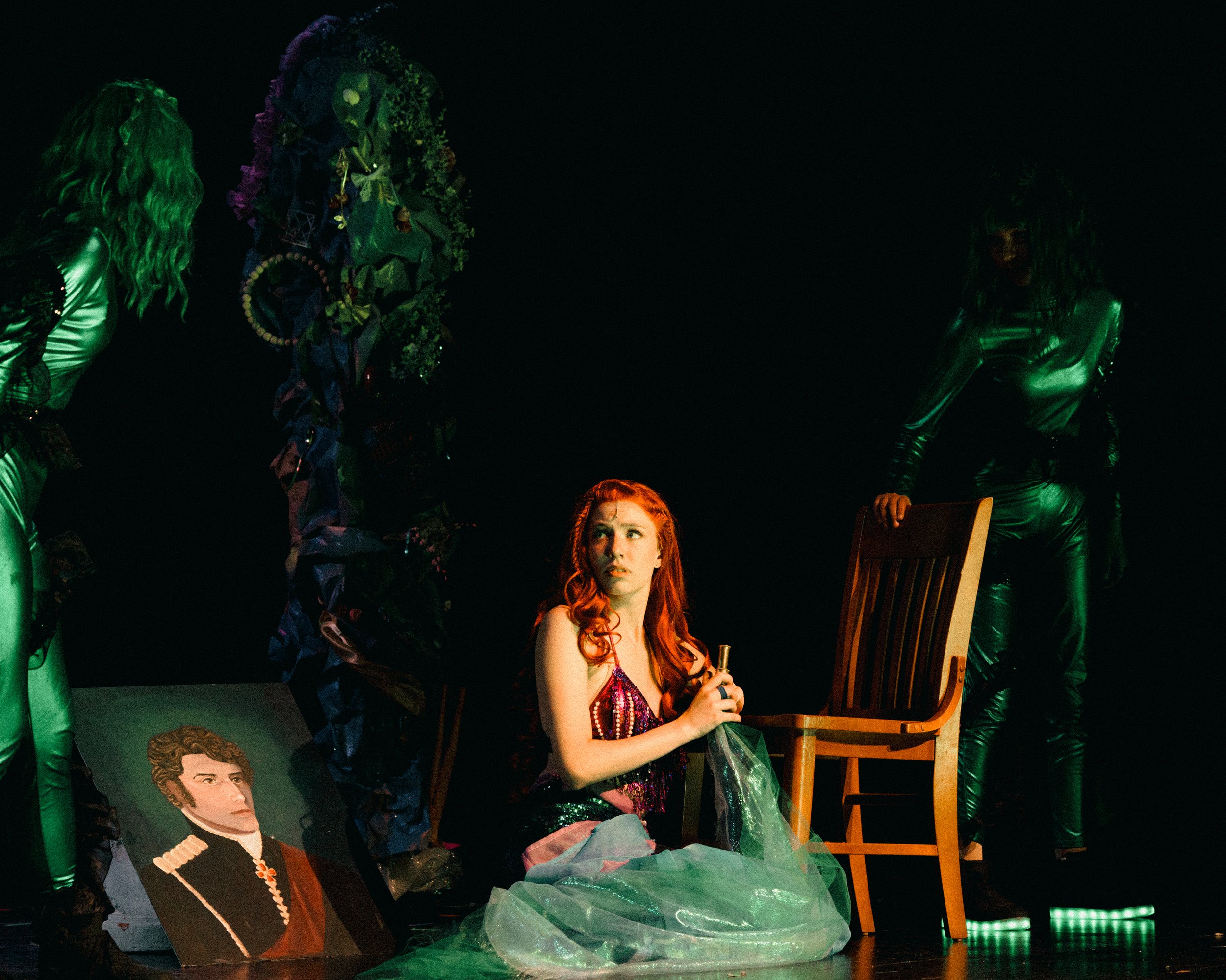 Stage scene with a woman sitting on the floor with red hair and a purple dress, surrounded by green lighting. There are two women in shiny outfits on either side, a chair, a framed portrait of a man, and a decorated large blue and purple arch behind 