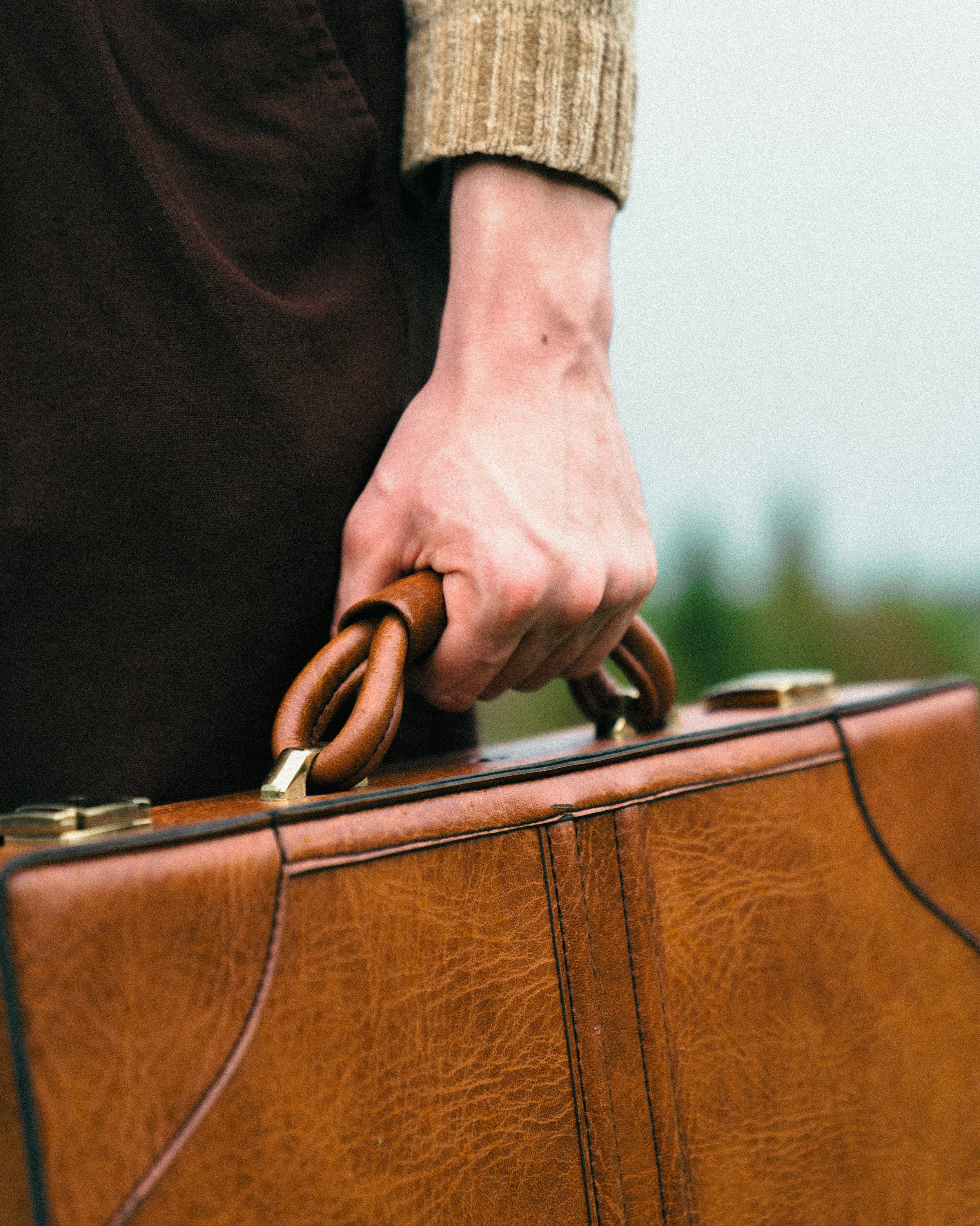 Close-up of a person holding a vintage brown leather briefcase with a wooden handle, wearing a beige sweater and dark pants.