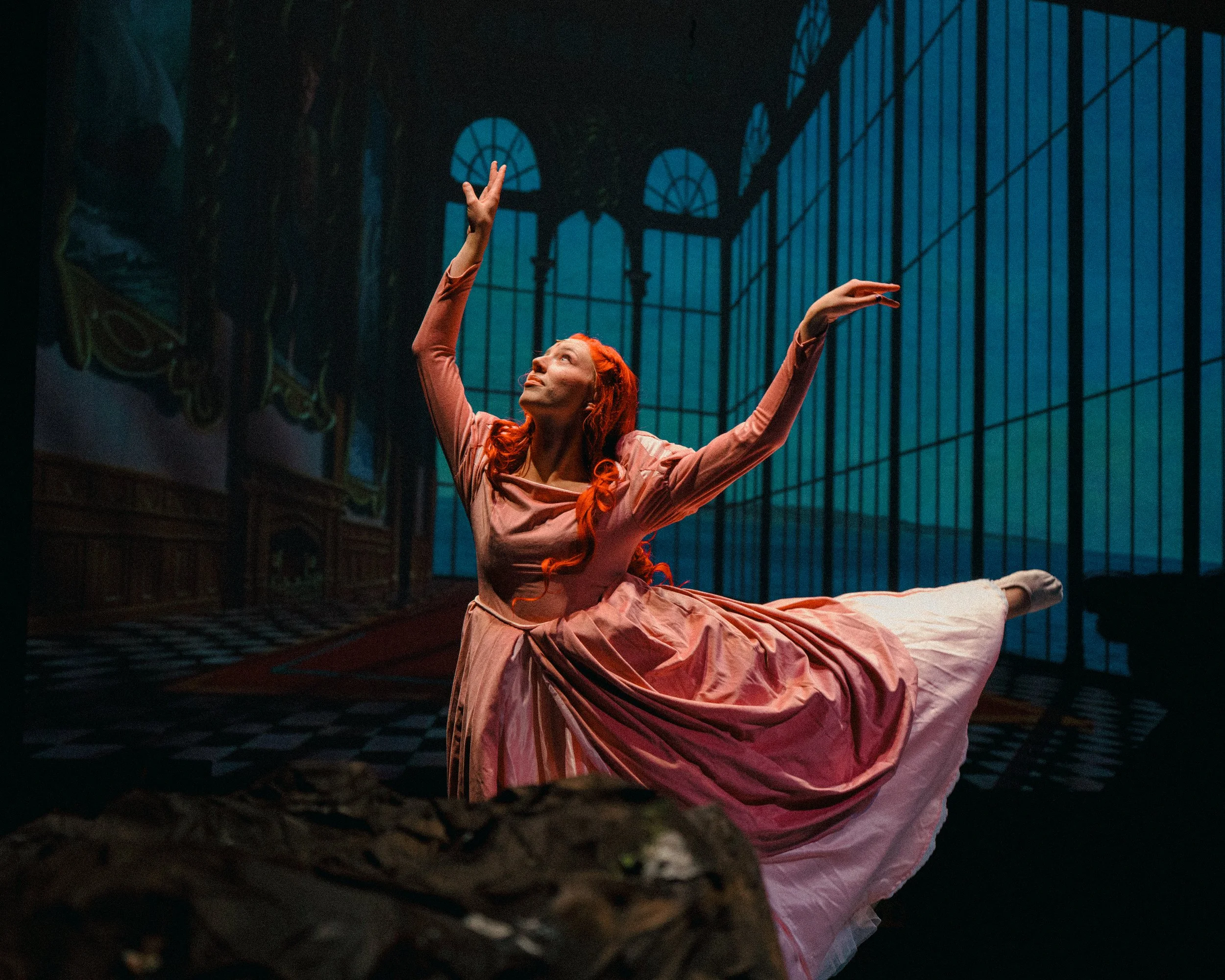 A woman ballet dancer with red hair performing a dance pose on stage, with a painted background of a large windowed conservatory.