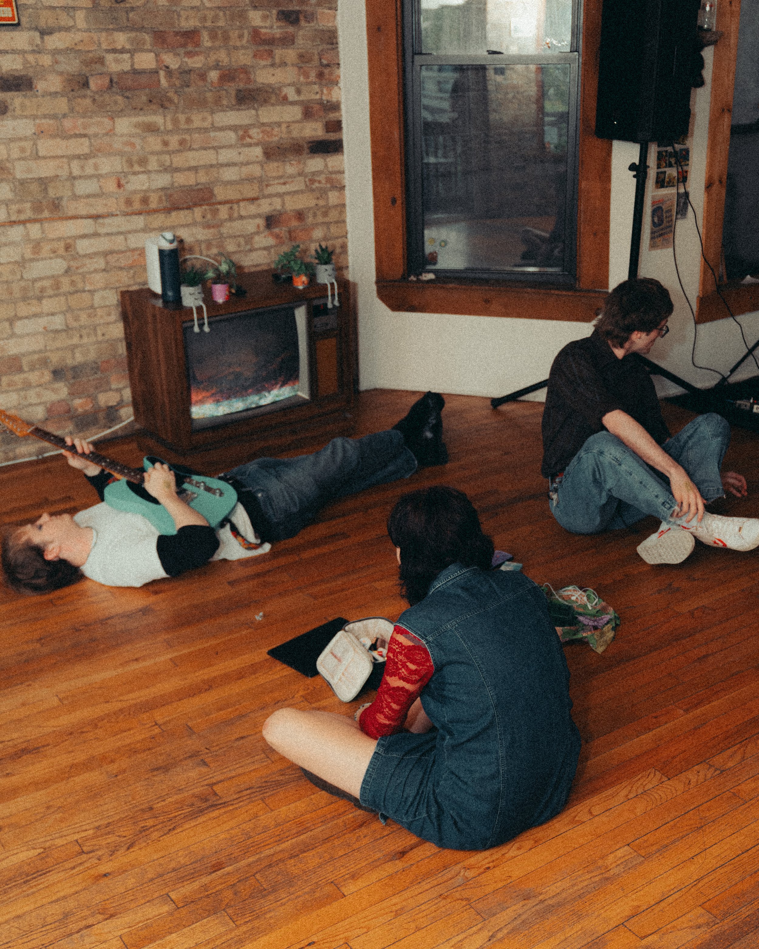 Three people and a dog in a cozy living room with brick and wood walls, one person lying on the floor playing an electric guitar, another sitting cross-legged looking at a phone, and a third sitting on the floor near a window. There is a small electr