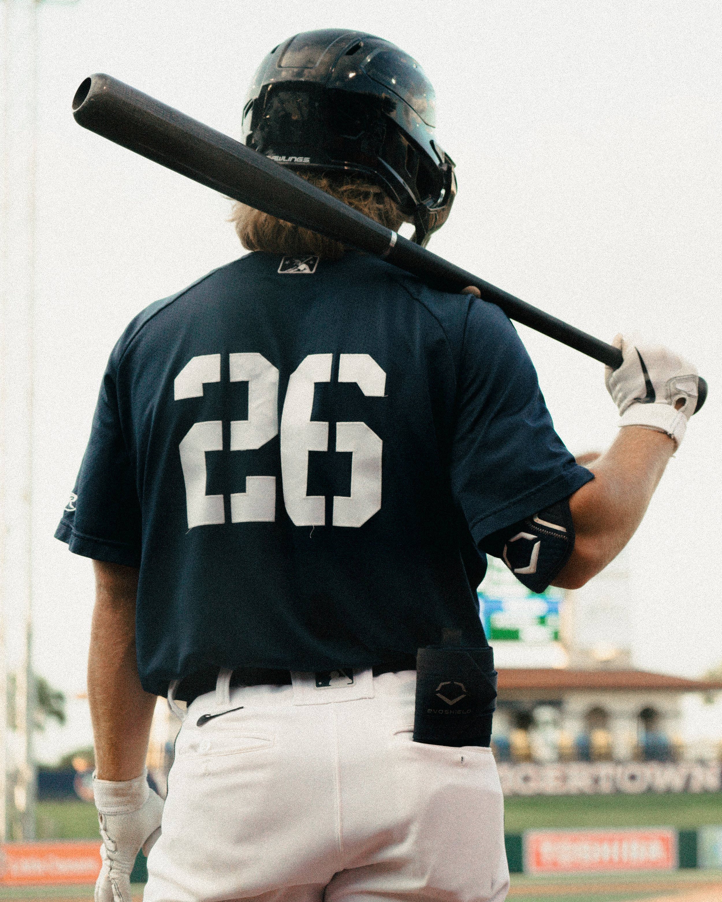 Back view of a baseball player wearing a navy blue uniform with the number 26 on the back, holding a bat over his shoulder, and a helmet on his head.