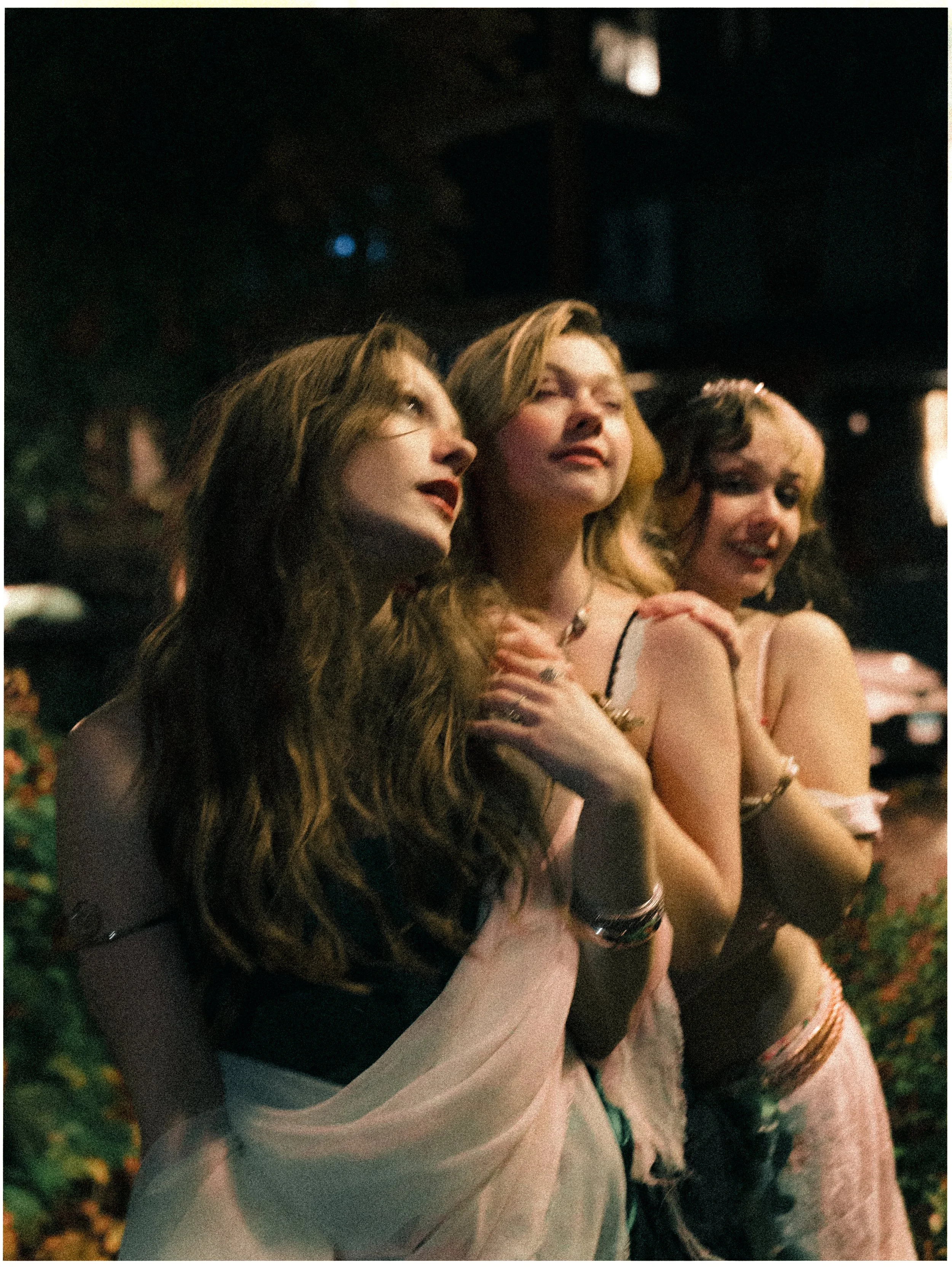 Three women with long hair standing close together at night, with one woman in the middle touching her shoulder, all wearing light-colored outfits and jewelry.