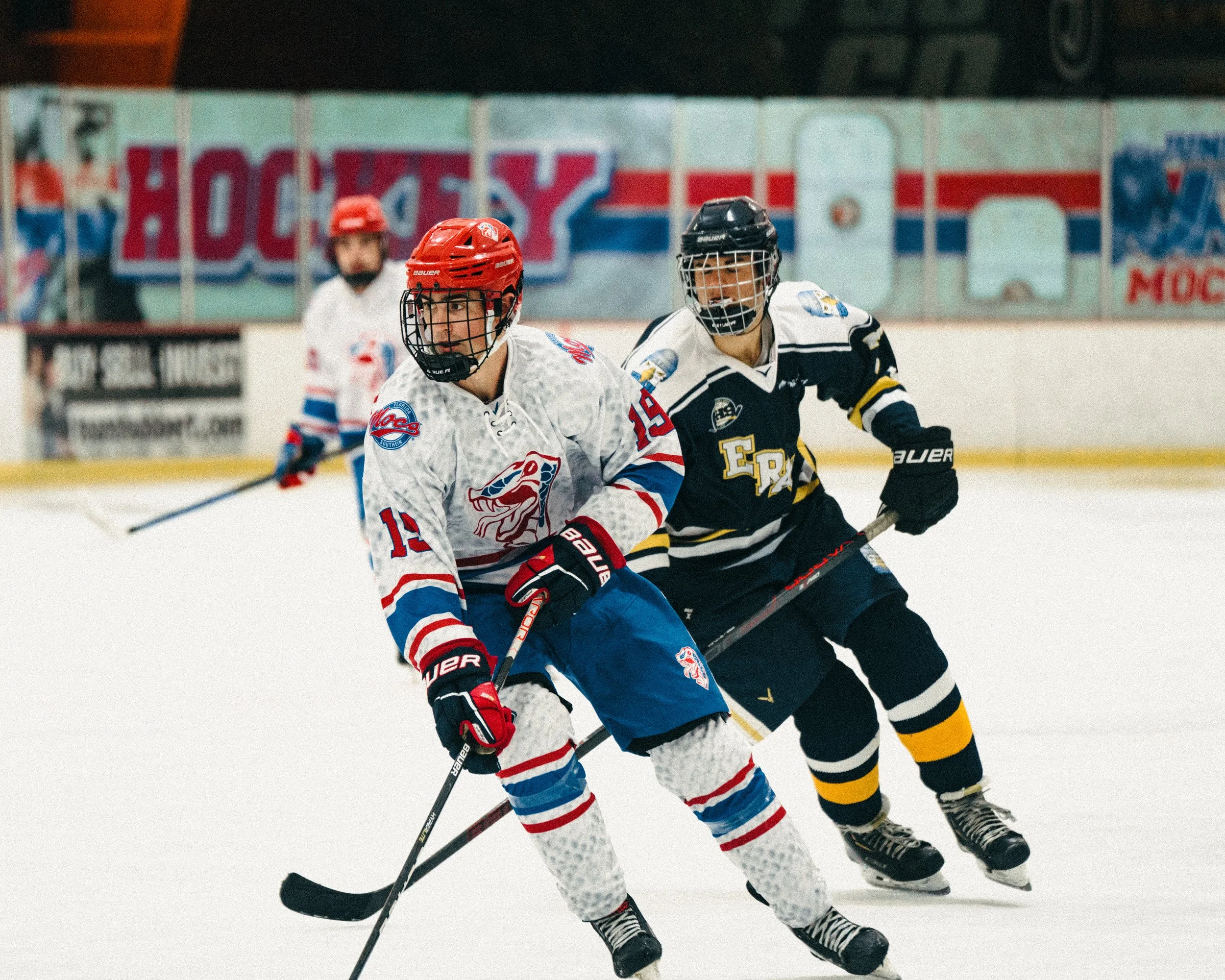 Two ice hockey players in action during a game, one in a white jersey with red and blue stripes and the other in a dark jersey with yellow and white accents, on ice rink. A third player is visible in the background.