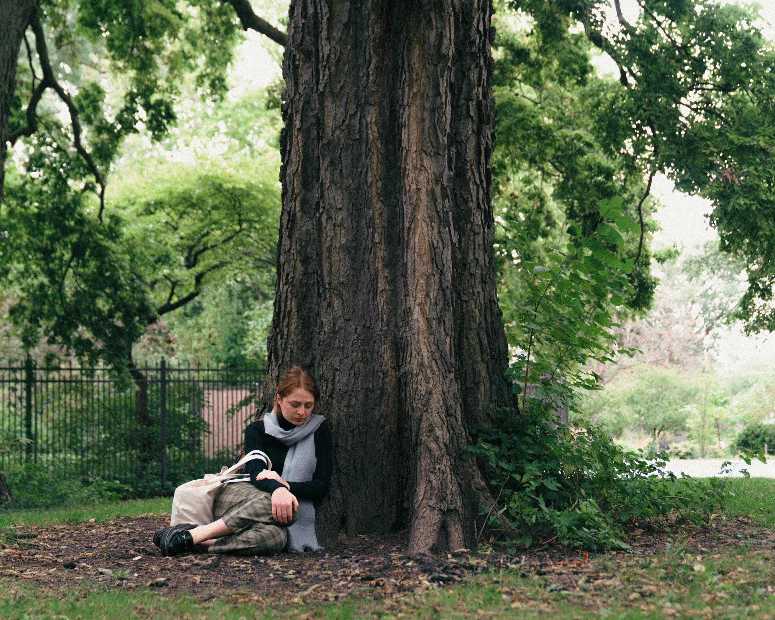 A young woman with red hair sitting on the ground, leaning against a large tree trunk in a park. She is wearing a black sweater, a light gray scarf, plaid pants, and black shoes. She appears to be looking down with a contemplative or sad expression. 