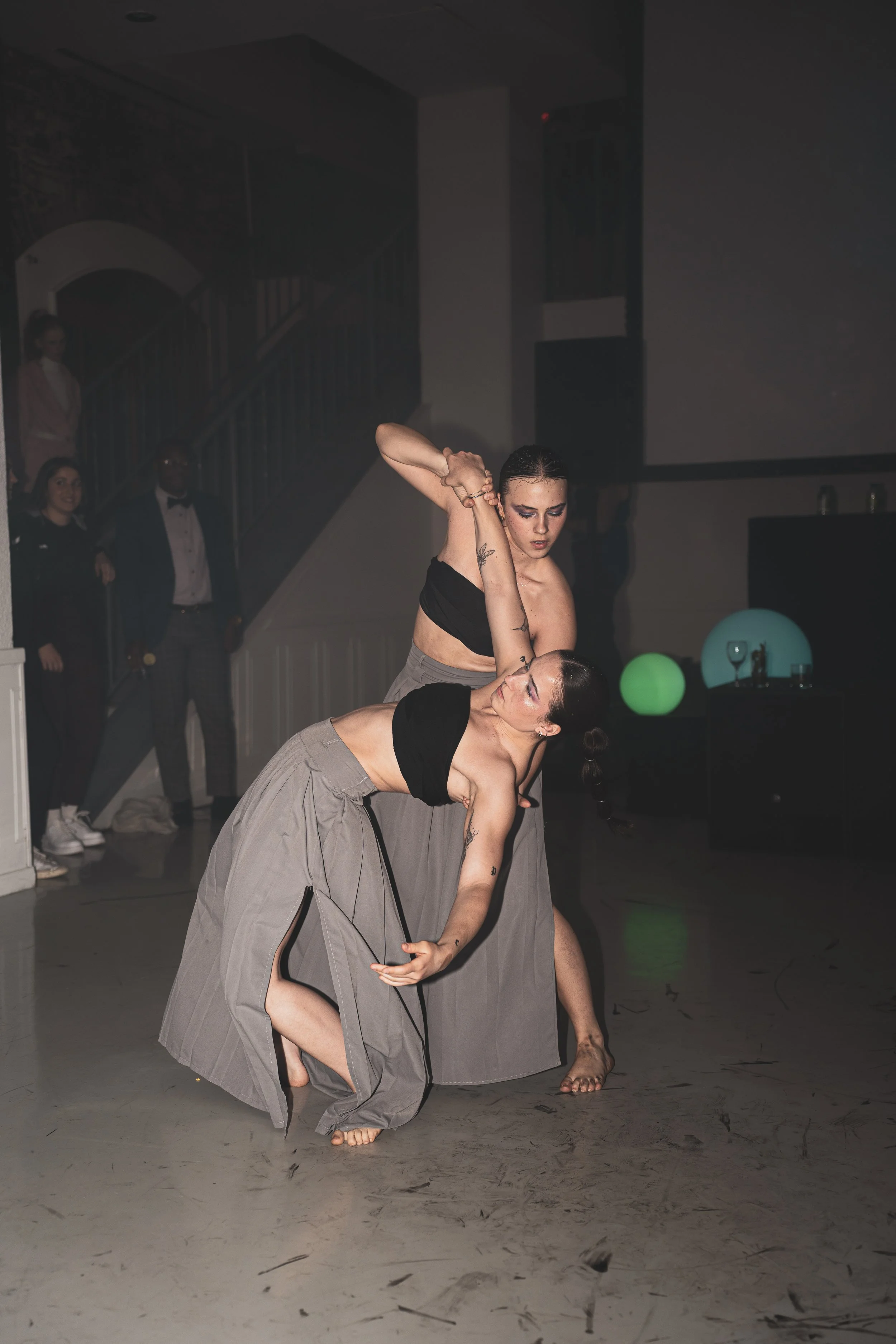 Deux femmes dansent en costume gris et noir lors d'une performance ou d'un spectacle en intérieur, avec quelques spectateurs en arrière-plan.