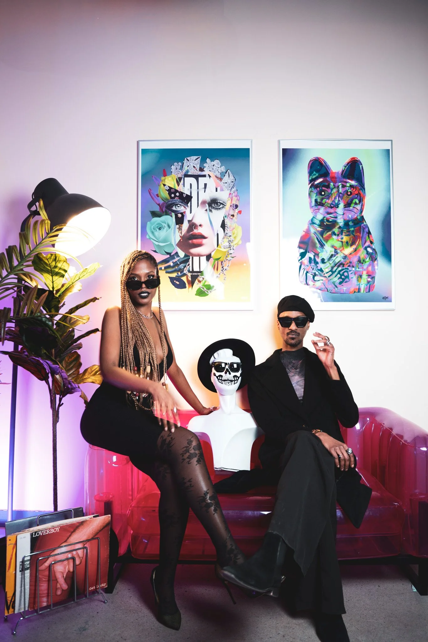 Deux personnes posent sur un canapé transparent rouge dans une pièce décorée, avec deux œuvres d'art colorées au mur, un mannequin avec un crâne maquillé, un lampadaire, une plante, et une collection de magazines.