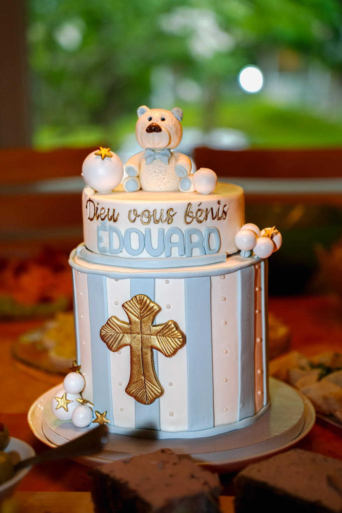 Gâteau de baptême blanc avec décoration dorée et bleue, figurine de ours en haut, croix dorée sur le devant, écrit en français 'Dieu vous bénit Edouard'