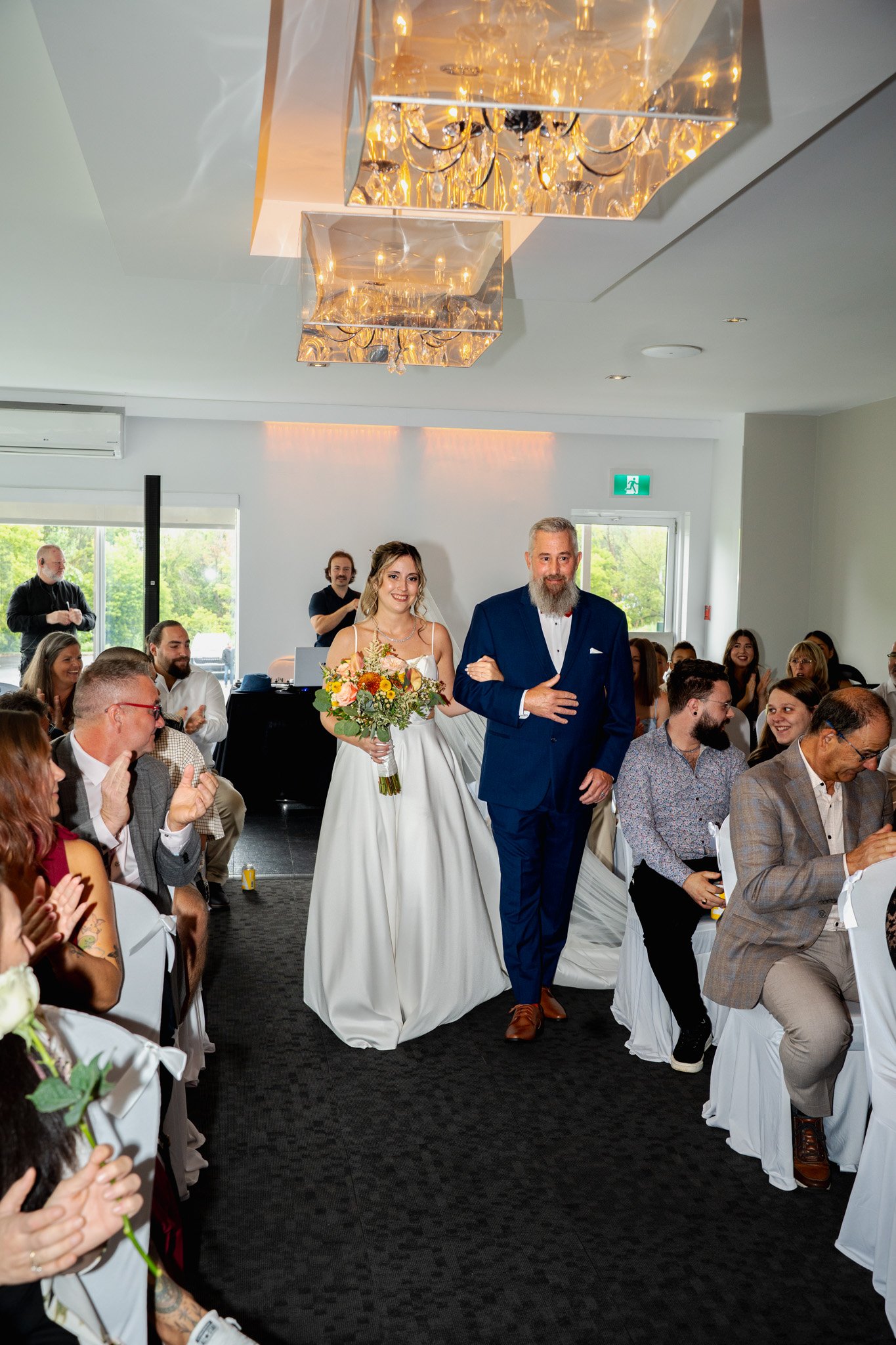Une mariée en robe blanche marche dans une salle de mariage, accompagnée d'un homme en costume bleu, entourée de convives applaudissant, dans une salle lumineuse avec un plafond décoré de lustres en cristal.