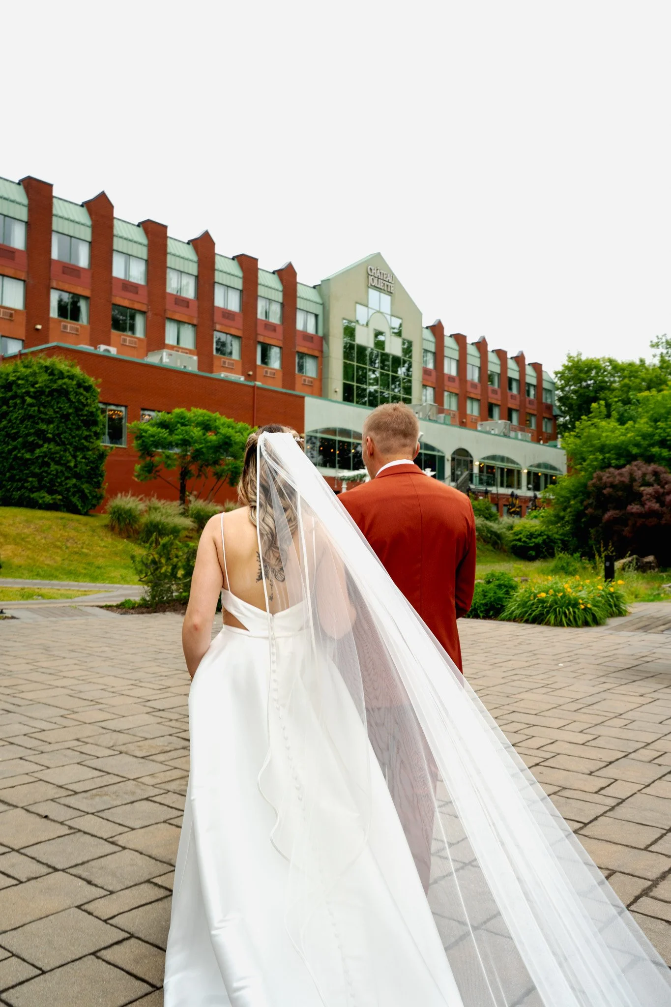 Un couple en robes de mariage marche vers un bâtiment nommé Château Olivette, dans un jardin, lors d'une cérémonie nuptiale
