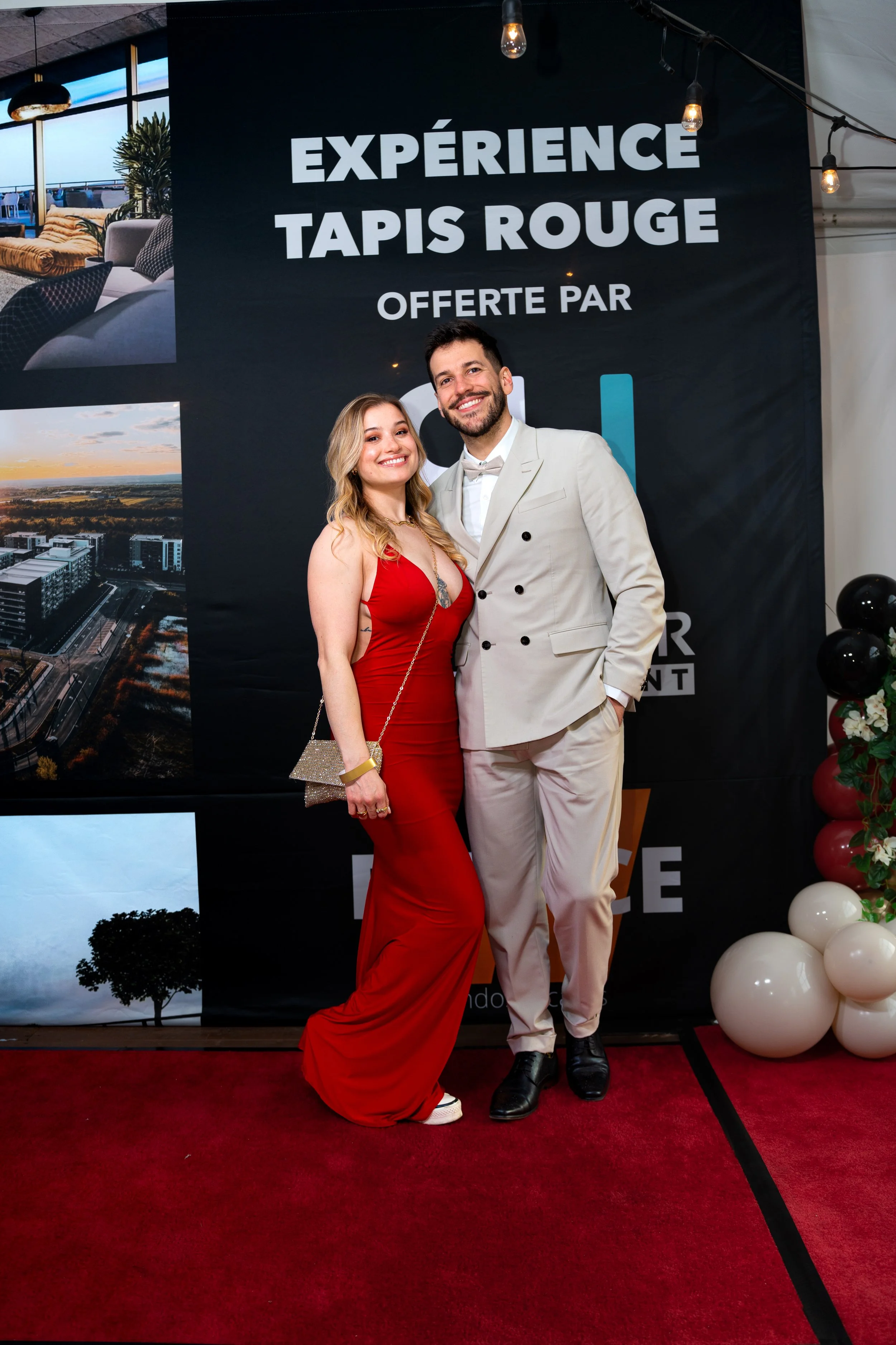 Un homme et une femme élégamment habillés posent ensemble lors d'un événement, devant un panneau noir avec du texte en français qui mentionne une expérience offerte par Tapis Rouge. La femme porte une robe rouge et le homme un costume blanc avec un nœud papillon.