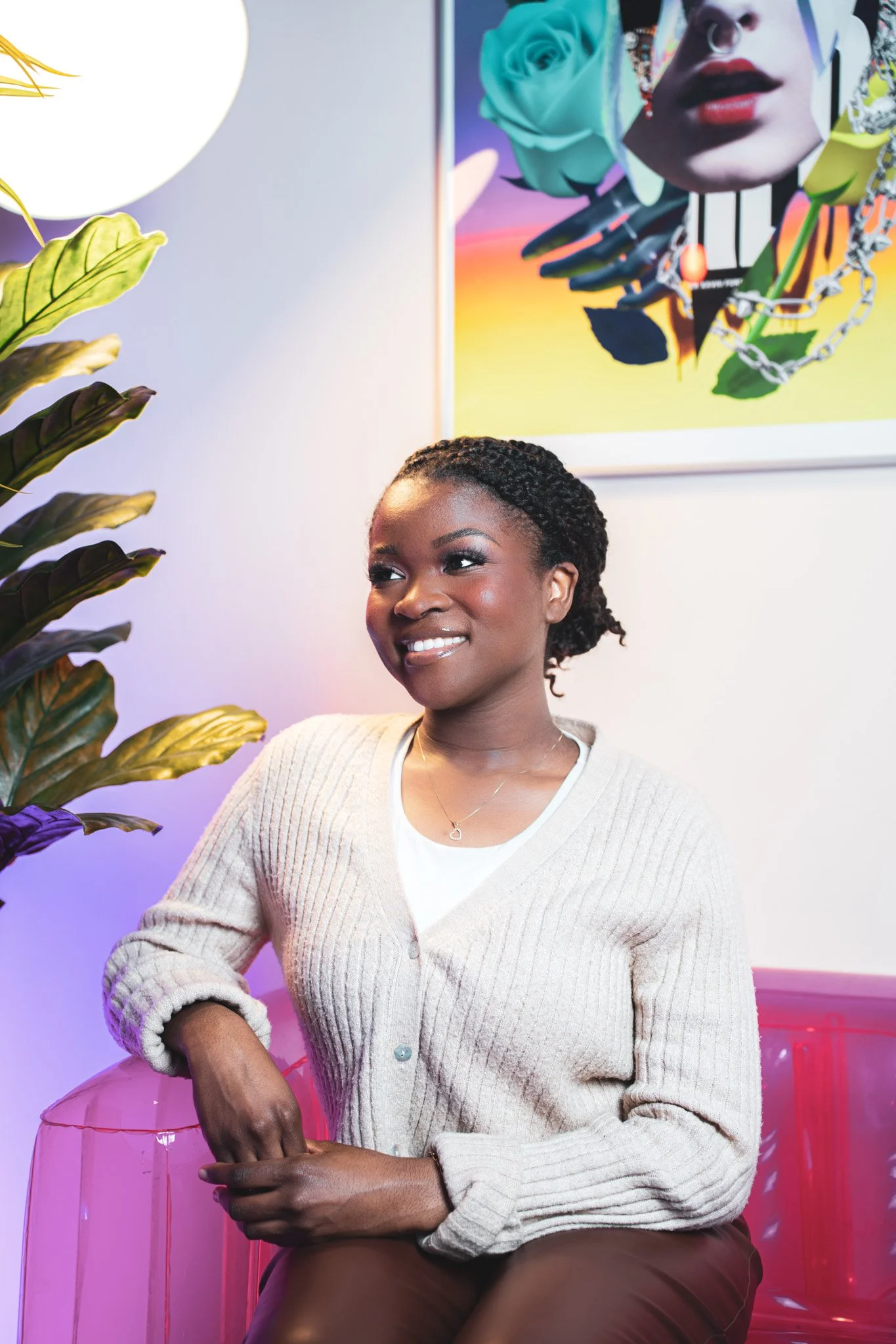 Jeune femme assise dans un espace coloré avec une peinture moderne en arrière-plan, portant un cardigan beige et un collier en forme de cœur.