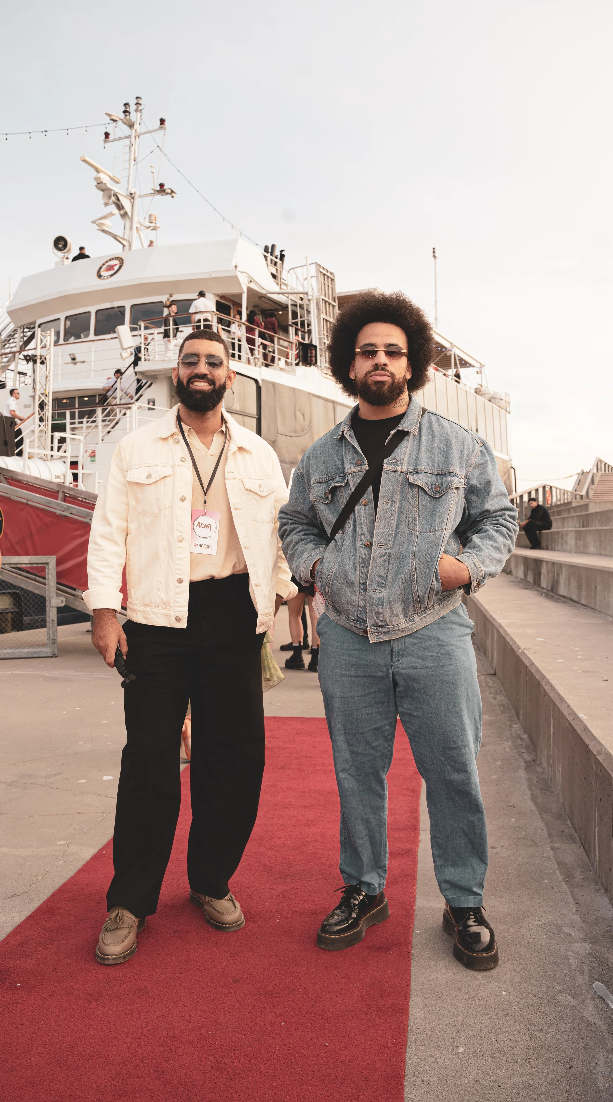 Deux hommes posent sur un tapis rouge près d'un bateau, un d'eux porte une veste blanche et l'autre une veste en denim, avec un ciel clair en arrière-plan.