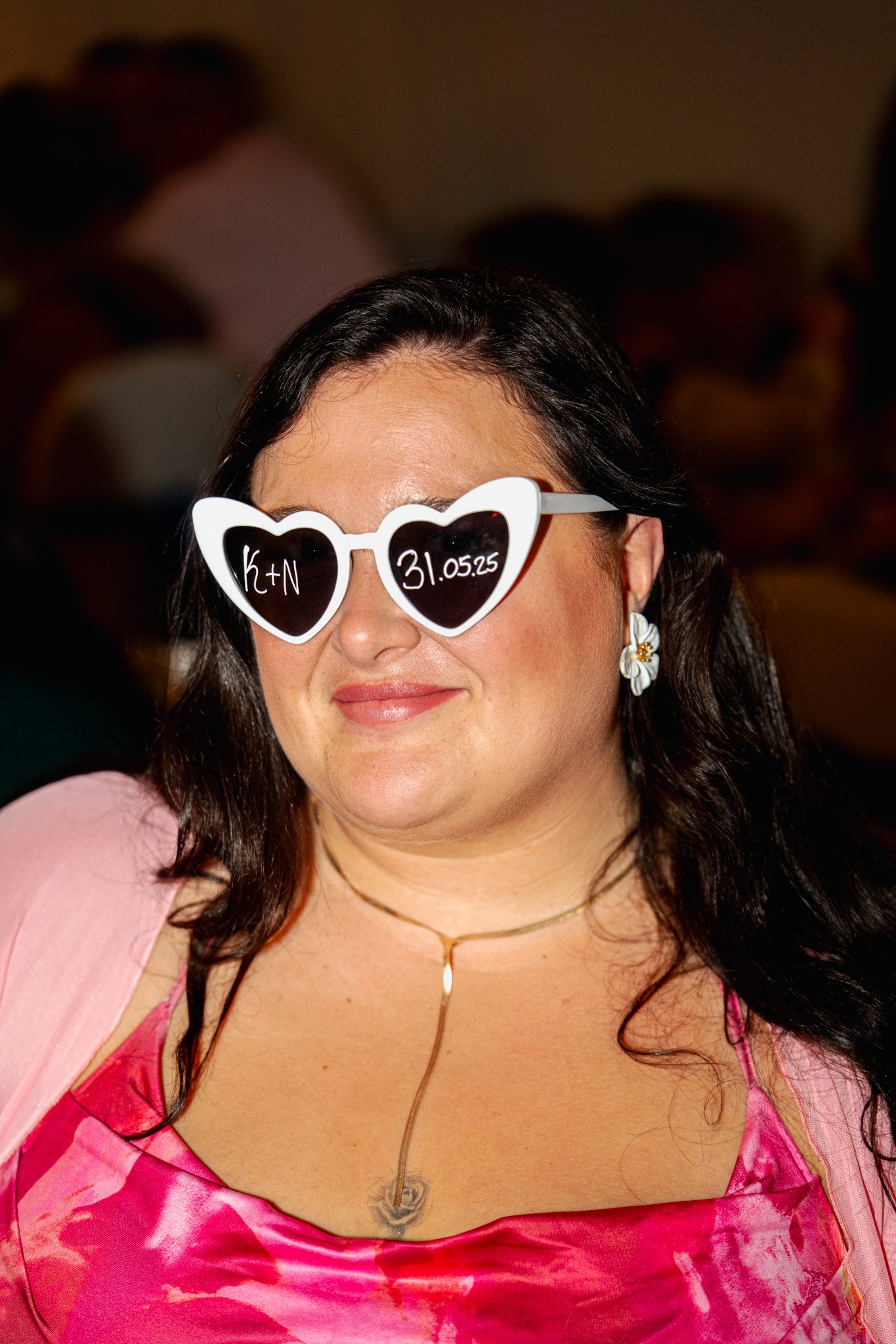 Femme portant des lunettes de soleil en forme de cœur avec écriture, portant une robe rouge et une veste rose, à une fête ou un événement.