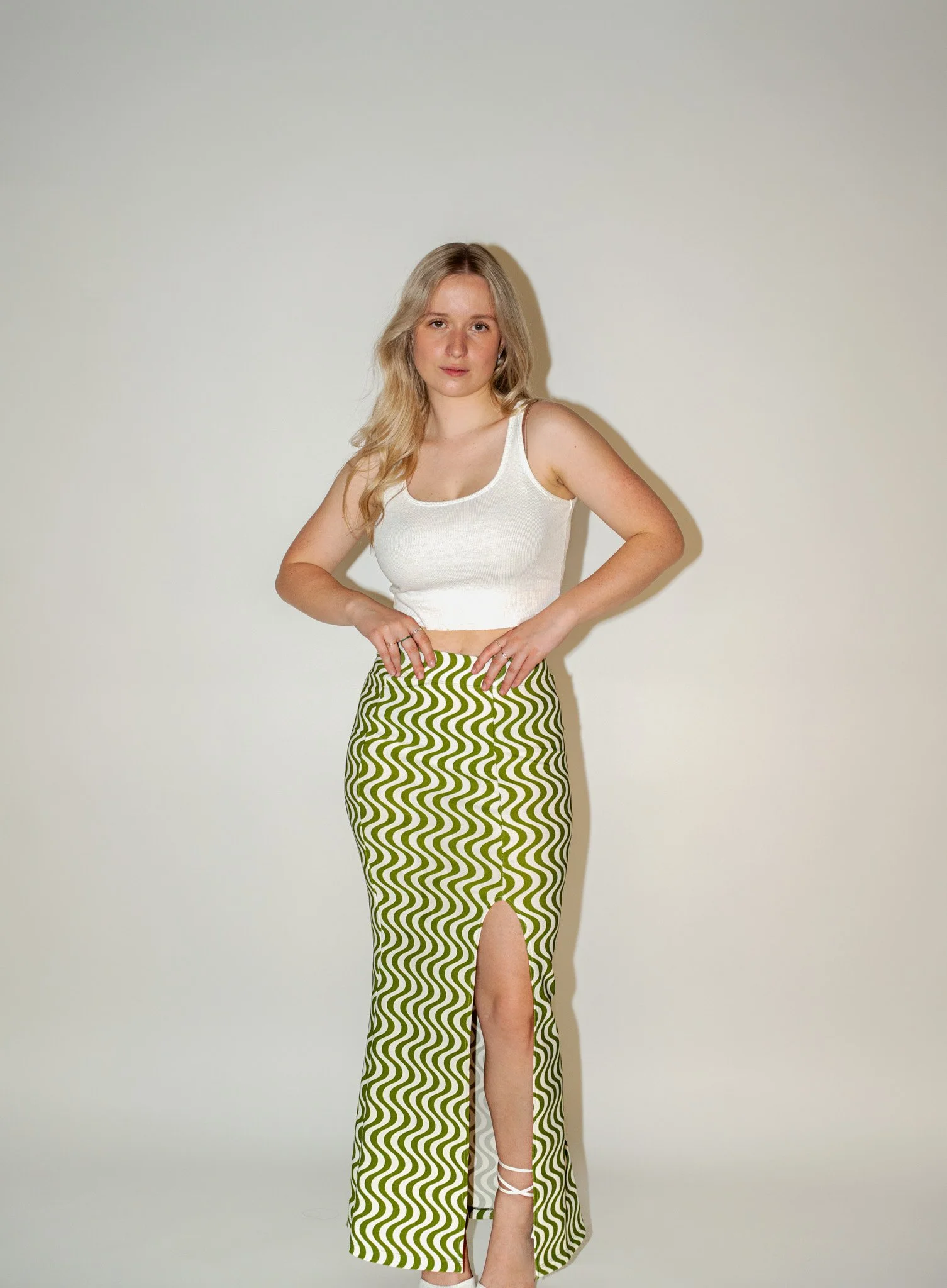 Jeune femme blonde avec une robe longue à jambes hautes avec un motif en zigzag vert et blanc, debout contre un mur beige clair.