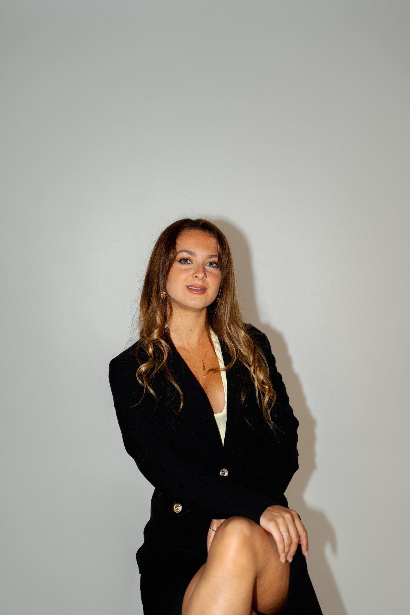 Une femme assise, portant un blazer noir, avec des cheveux longs ondulés, posant devant un mur blanc.