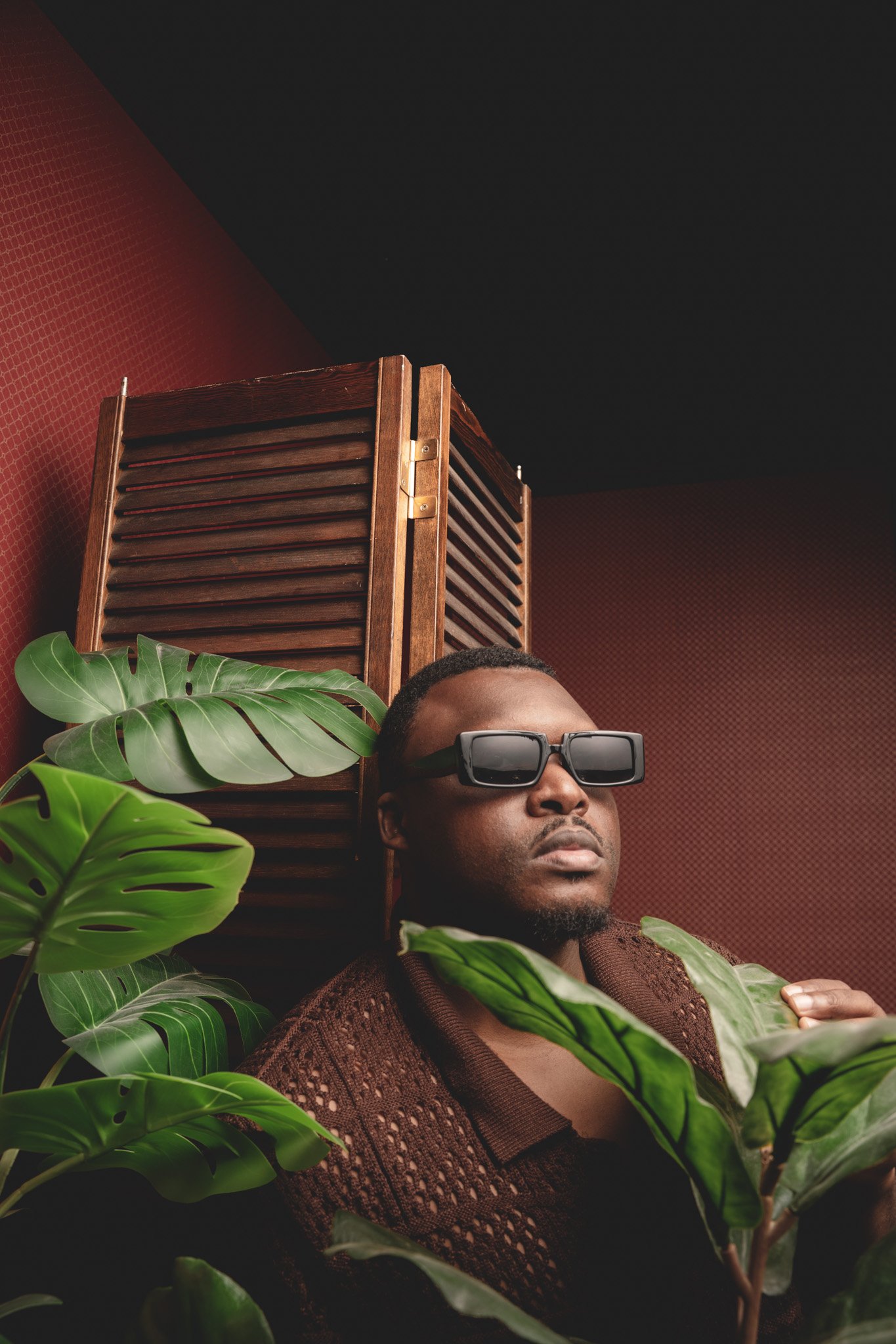 Un homme portant des lunettes de soleil noirs, entouré de plantes vertes, avec un mur rouge foncé et une cloison en bois derrière lui.