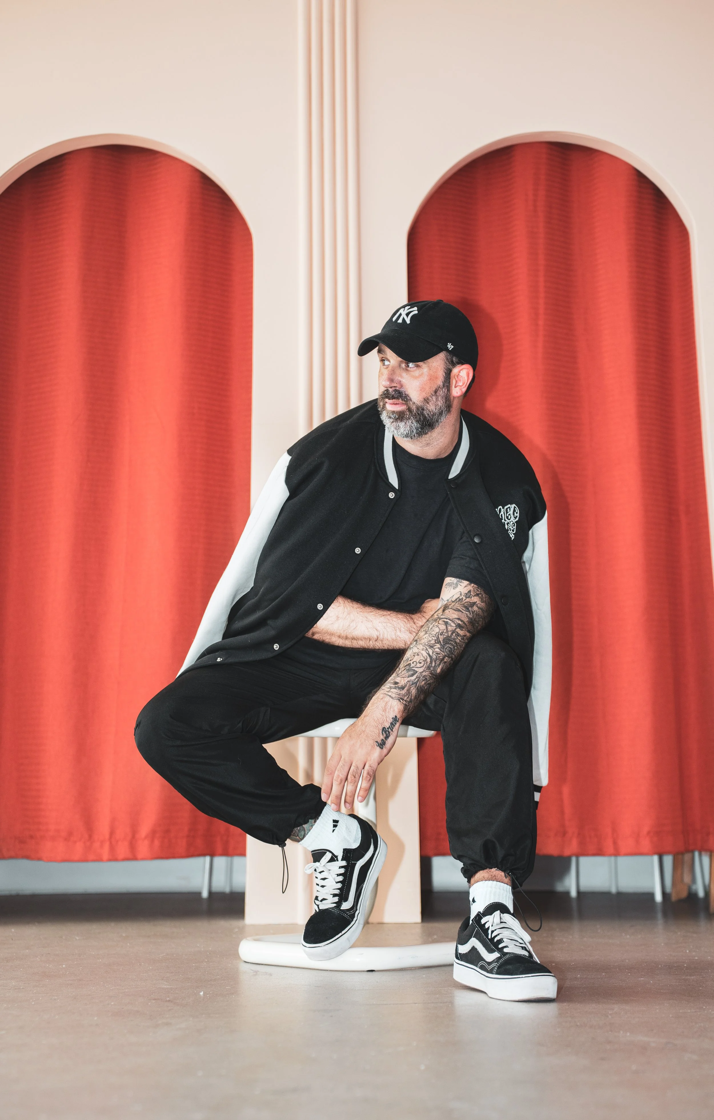 Un homme assis sur une chaise en face d'un drap rouge, portant un sweat à capuche, un casquette et des sneakers, avec un tatouage visible sur le bras.