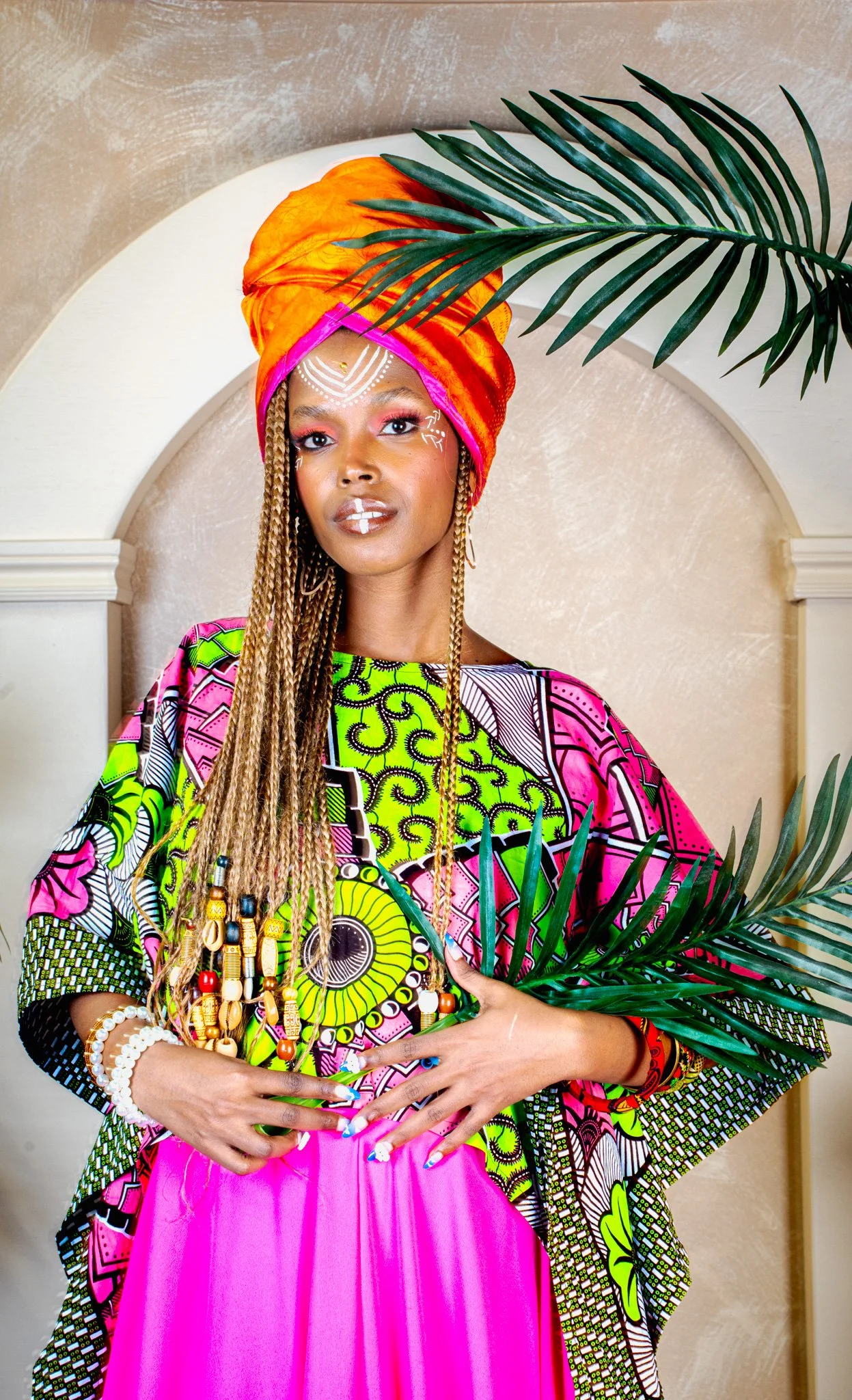 Jeune femme africaine portant un tissu coloré, un turban orange et rose, des bijoux, tenant des feuilles de palmier, avec un maquillage traditionnel lumineux.