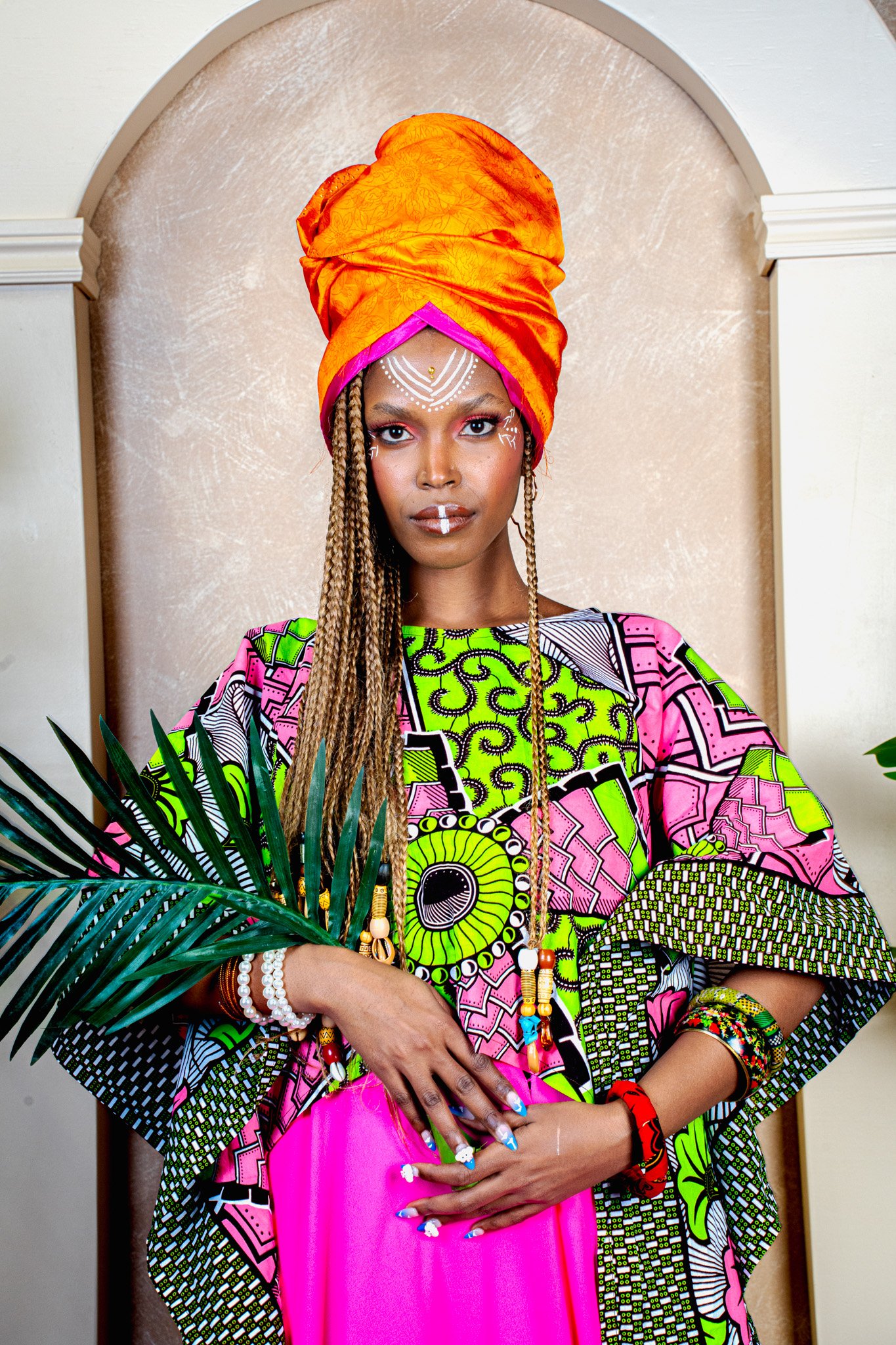 Femme portant un turban orange, vêtement coloré avec des motifs africains, tenant une feuille de palmier, portant des bijoux, avec un maquillage pastel et artistique.