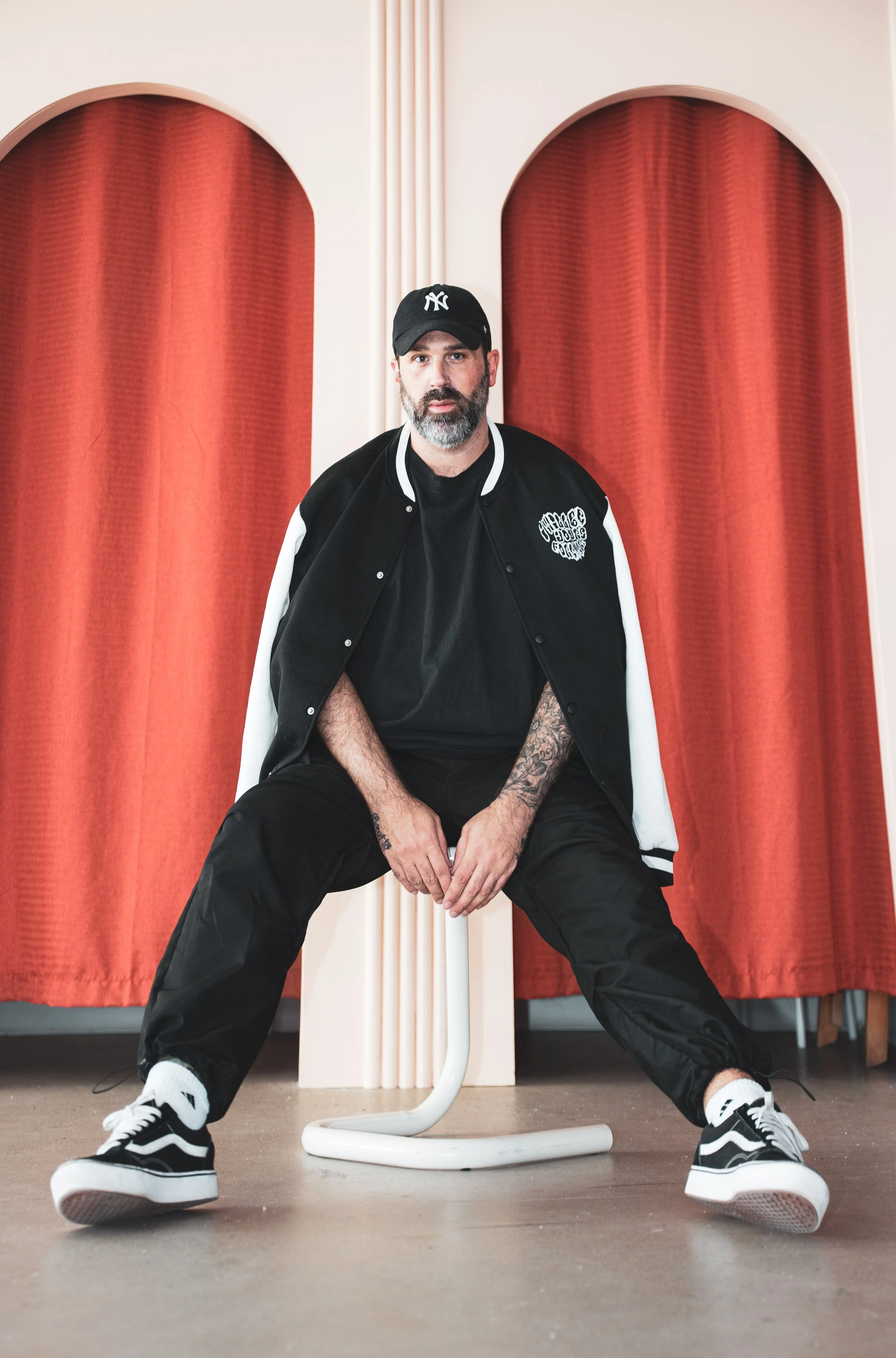 Un homme assis sur un tabouret, portant une veste de baseball noire et blanche, un t-shirt noir, un jean noir, une casquette noire et des chaussures de skate noires et blanches, devant un fond avec des rideaux rouges et un décor beige.