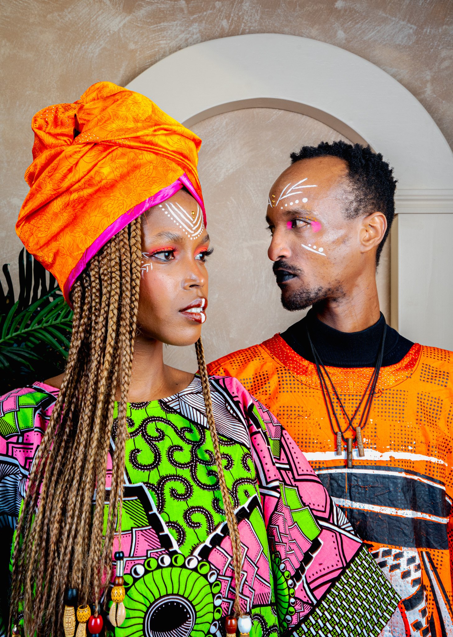 Deux personnes portant des vêtements et un maquillage traditionnels colorés, se regardant intensément, dans un intérieur avec mur beige et décor en forme d'arc blanc.