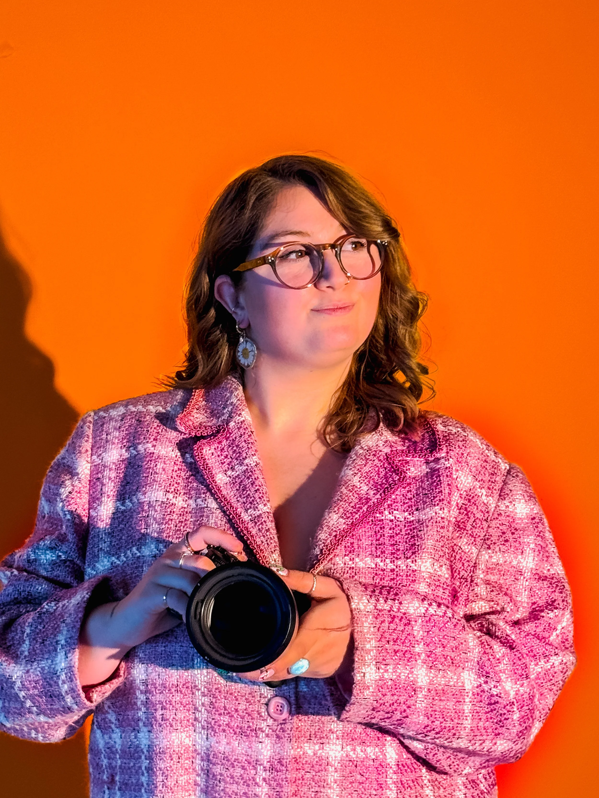 Une femme avec des lunettes, tenant un appareil photo, portant un blazer rose vif, avec un fond orange.