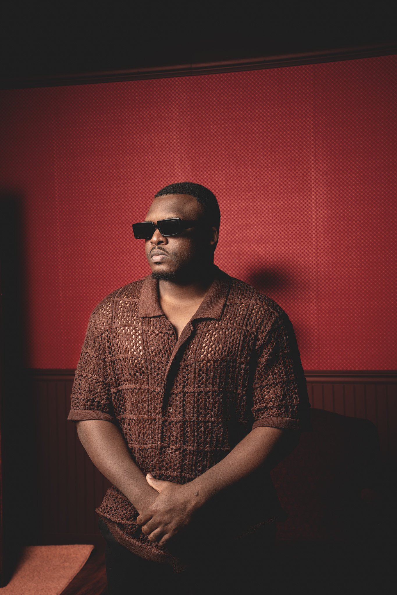 Homme portant des lunettes de soleil sombres, cheminée dans une pièce avec mur rouge et panneau en bois, portant un pull marron.