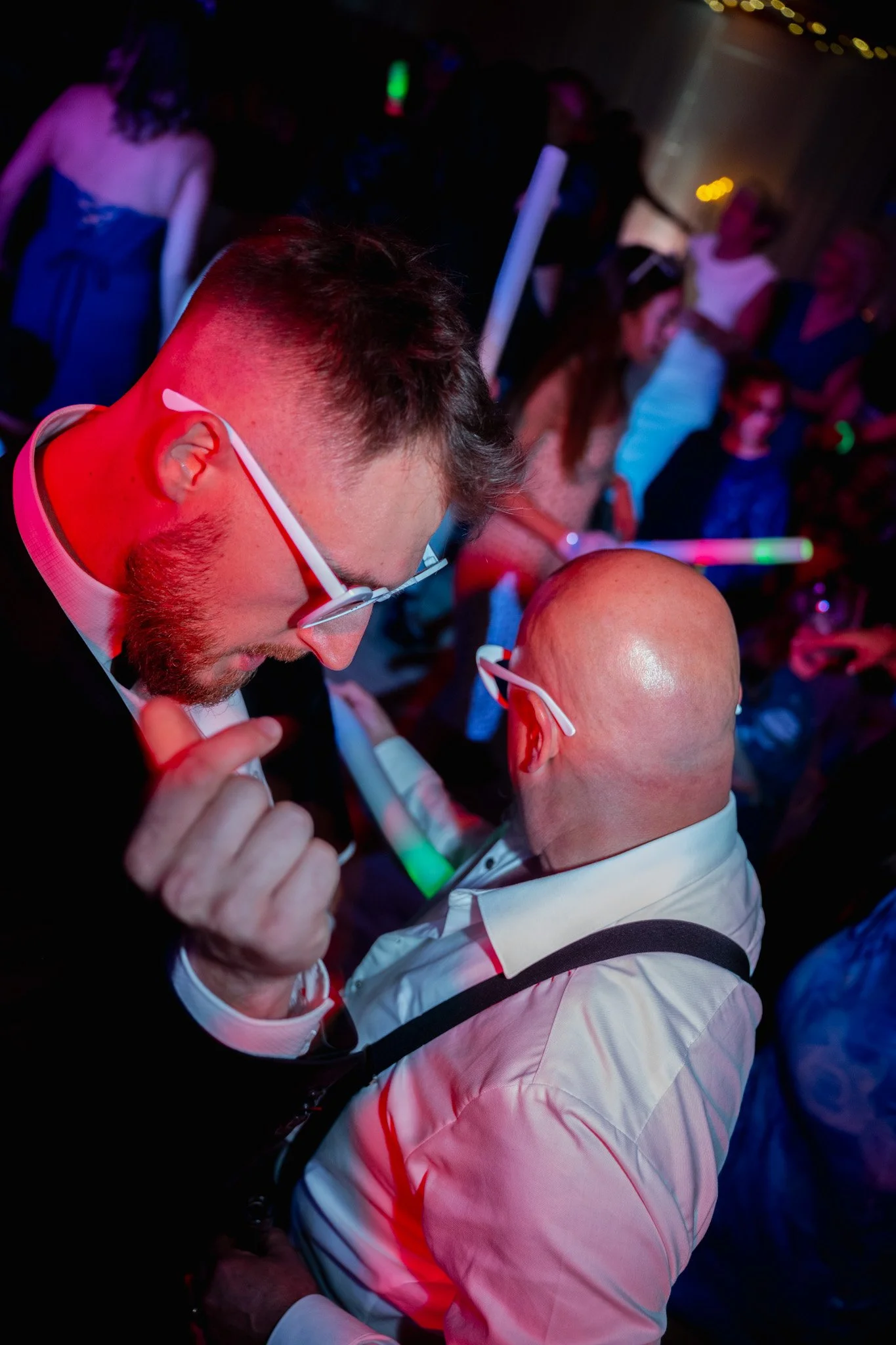 Deux hommes portant des lunettes blanches dansent lors d'une fête ou une soirée, entourés d'autres personnes, dans une ambiance colorée et animée.