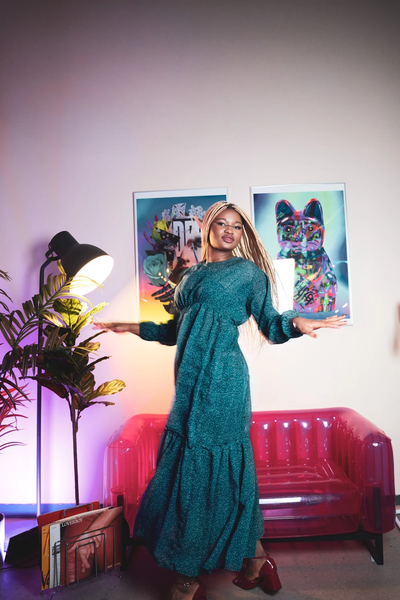 Une femme pose dans un intérieur modern avec un canapé rouge transparent, une lampe et deux affiches d'art contemporain au mur, portant une robe longue verte et des chaussures à talons bordeaux.