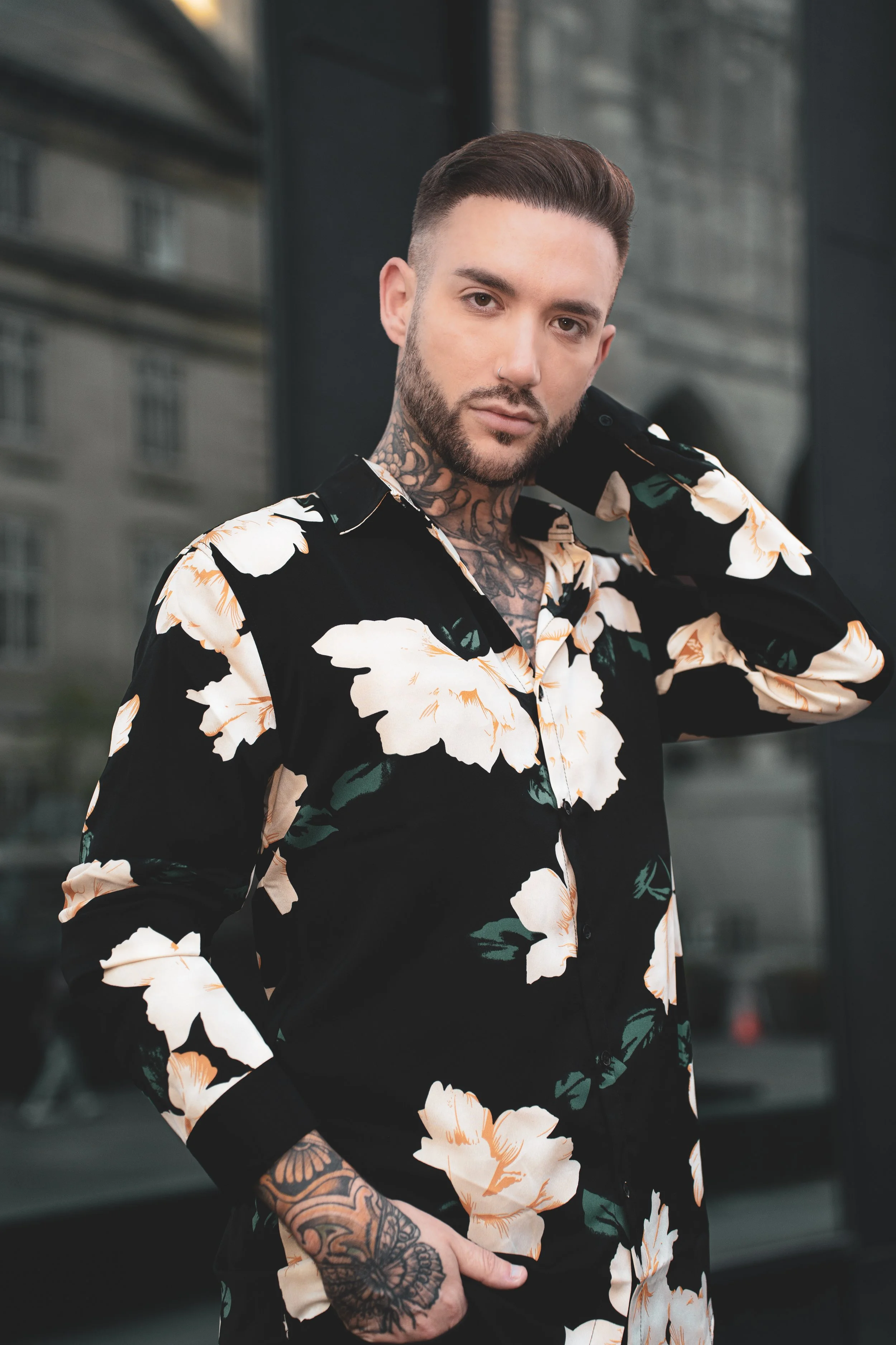 Homme avec des tatouages portant une chemise noire à fleurs blanches, posant devant un bâtiment urbain.