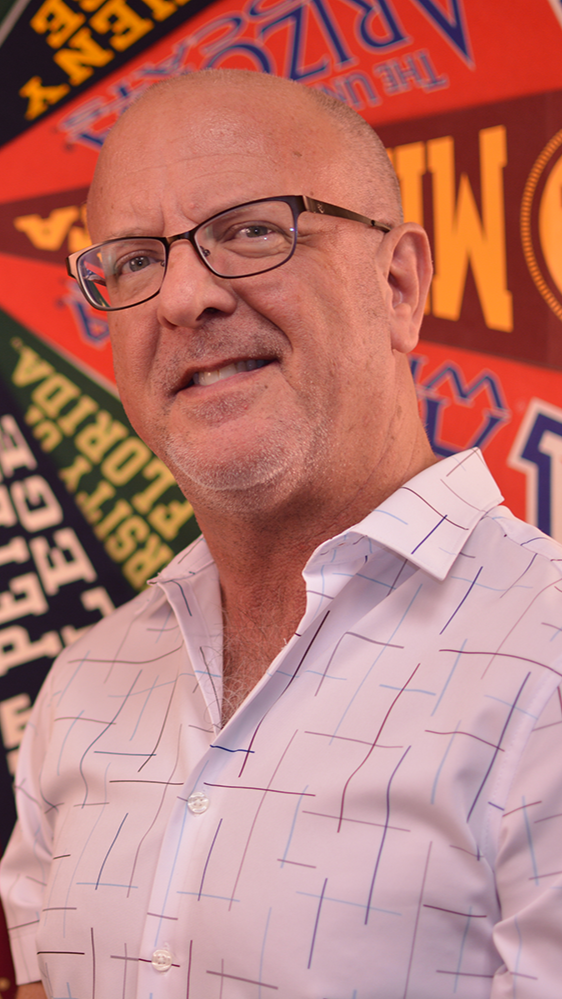 A man with glasses and a bald head smiling, wearing a white dress shirt with thin, colorful grid patterns, standing in front of a colorful, patterned background.