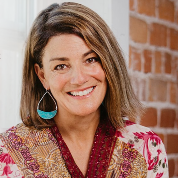 A woman with shoulder-length brown hair smiling, wearing a colorful patterned blouse and a teal earring, with a brick wall background.
