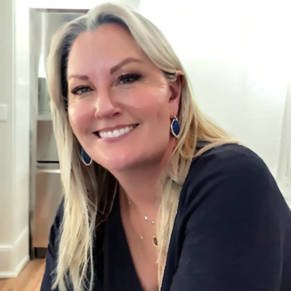 A smiling woman with blonde hair wearing a black top and blue earrings, indoors with a refrigerator in the background.