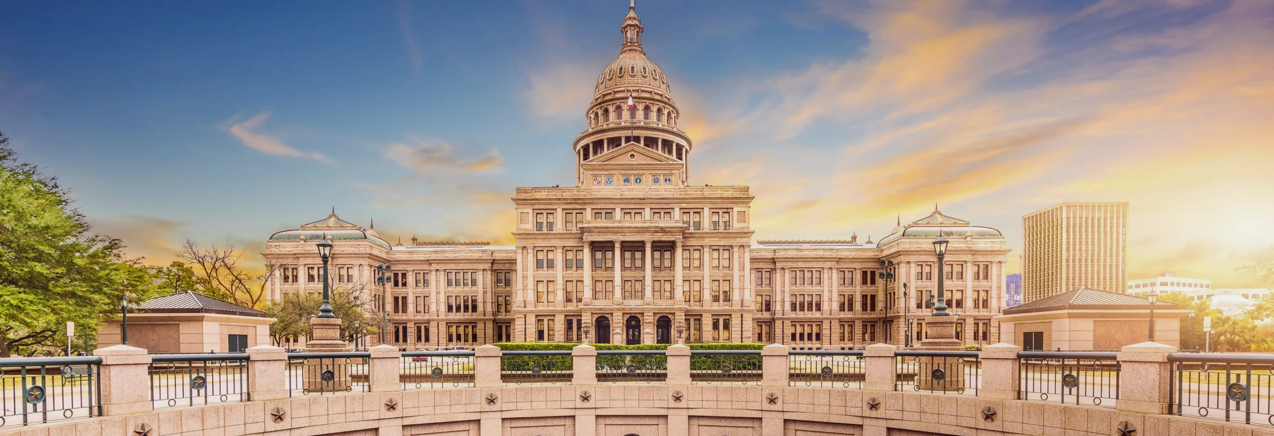 The Texas State Capitol building at sunset with a cloudy sky, trees on the left, and a city skyscraper on the right.