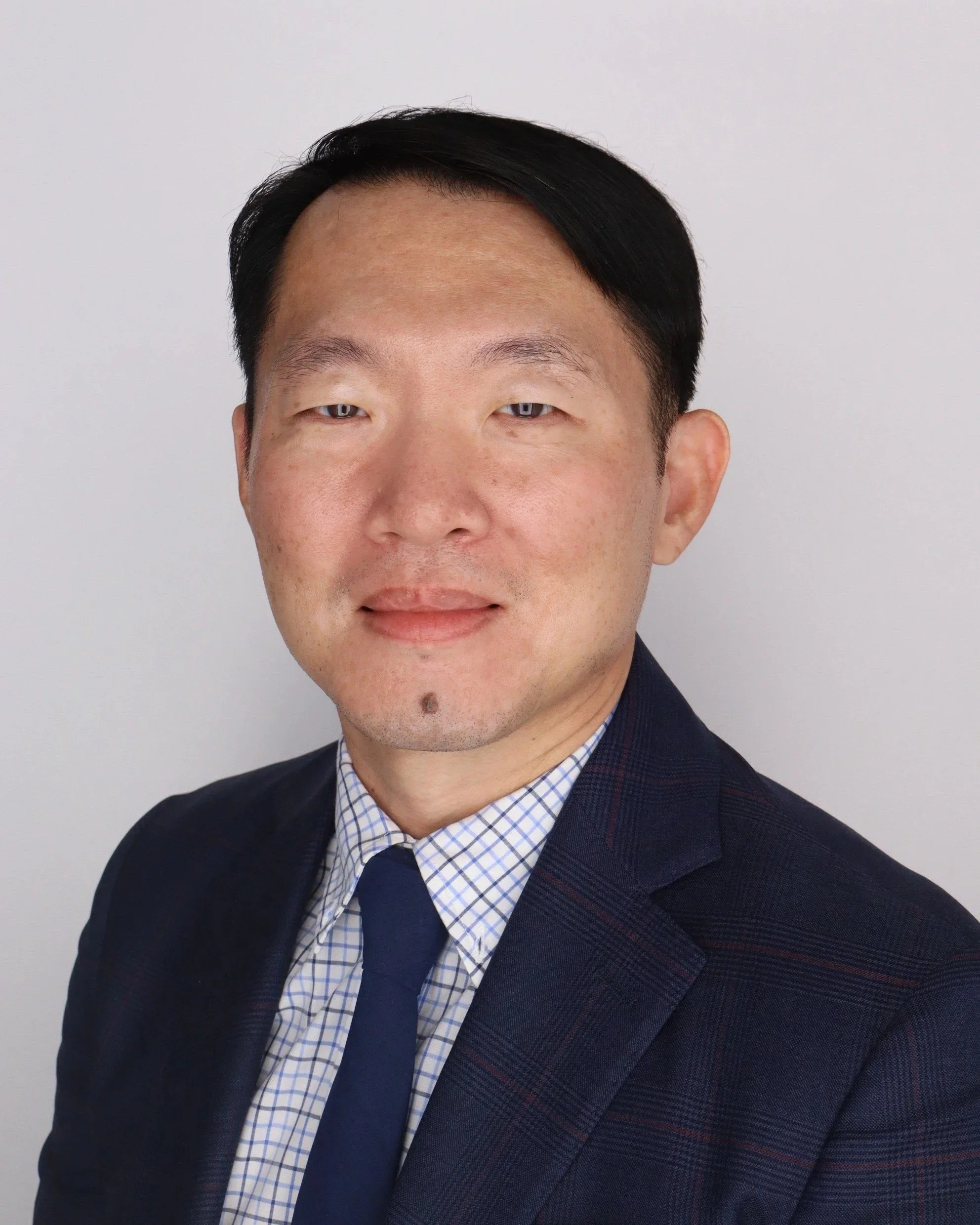 Close-up photo of an Asian man wearing a dark suit and blue checkered shirt, smiling slightly, against a plain white background.