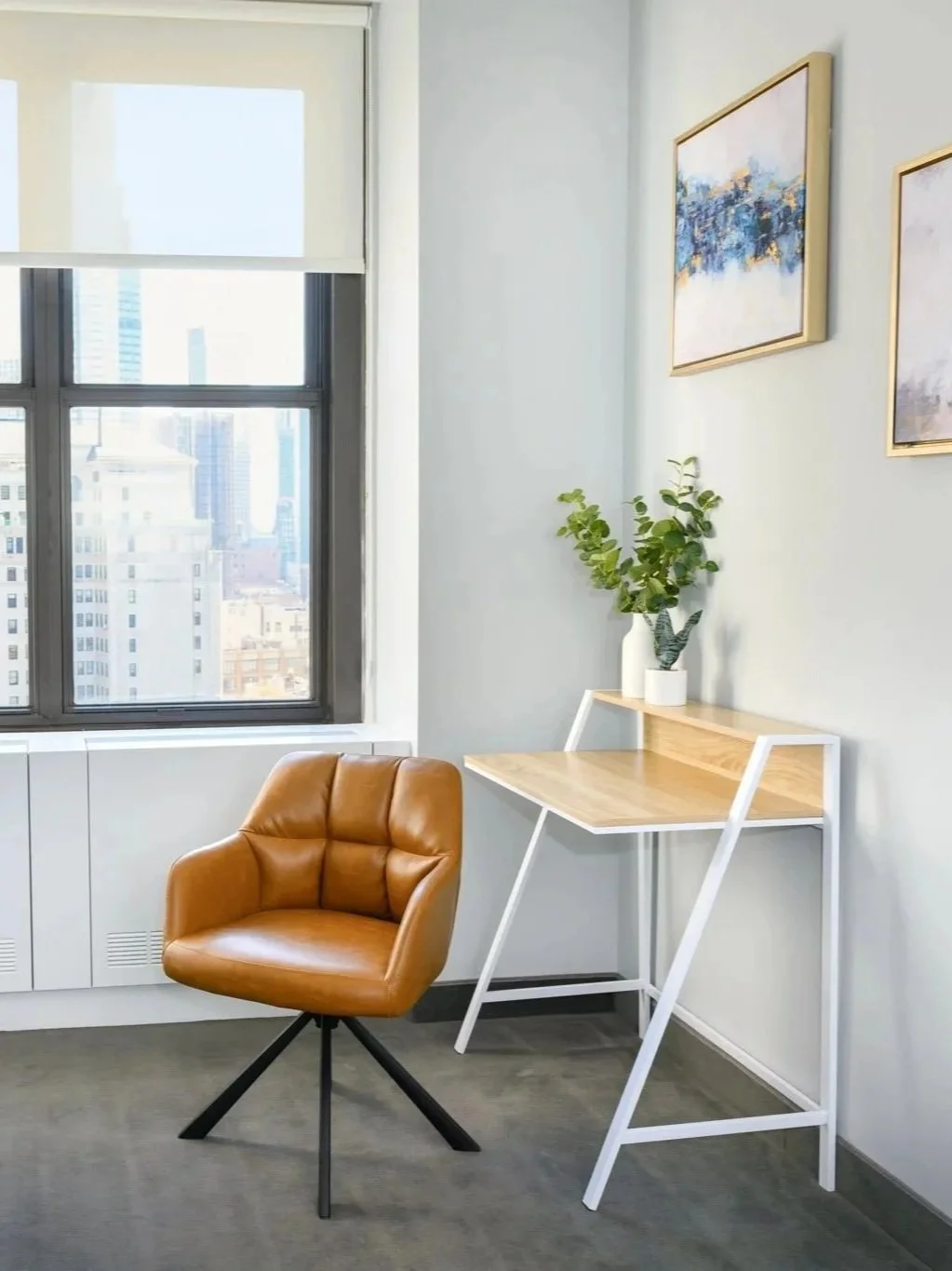 A corner of an office room with a window showing a cityscape view, a tan leather swivel chair, a small wooden desk with white metal legs, a white vase with green leafy plants, and two abstract framed paintings on the wall.
