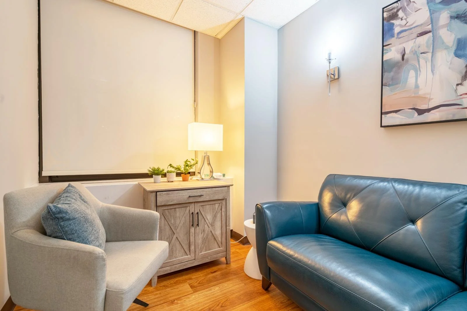 A cozy living room corner with a light grey armchair, a small wooden cabinet with a lamp and potted plants on top, and a dark blue leather sofa. There's a large window with a closed white blind, and a colorful abstract painting hanging on the wall. The room has wooden flooring and warm lighting.