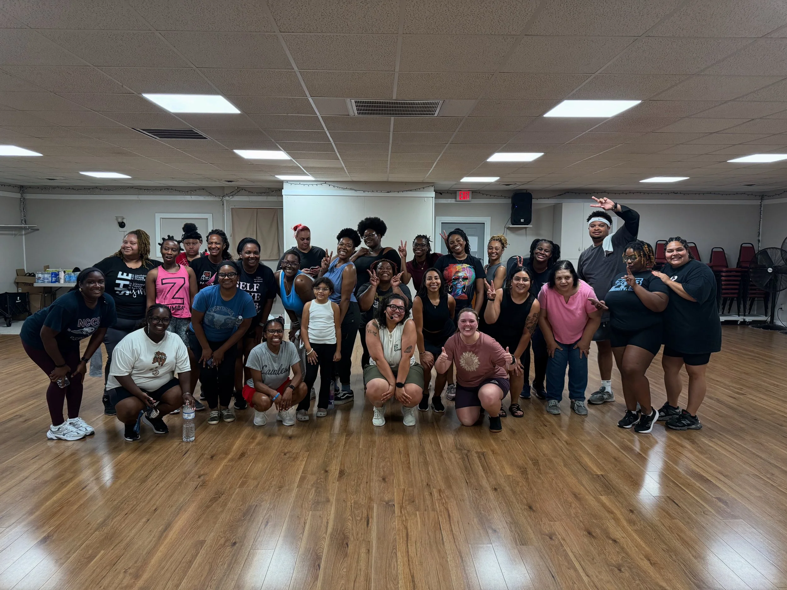 Group of smiling people posing for a photo in an indoor hall with wooden floors and a white wall background.