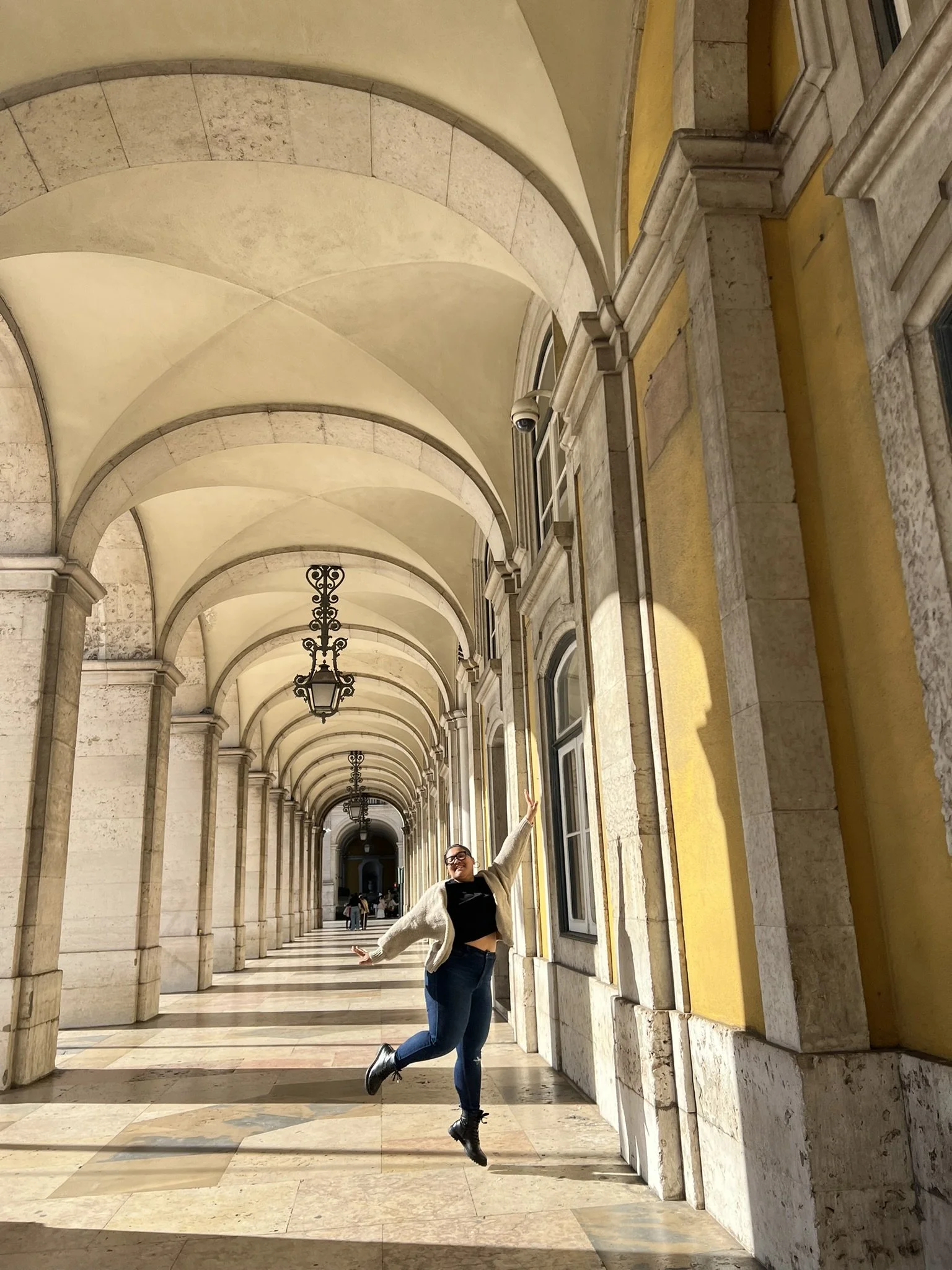 A young woman jumping joyfully in a historic corridor with arches and chandeliers, sunlight streaming through the windows.