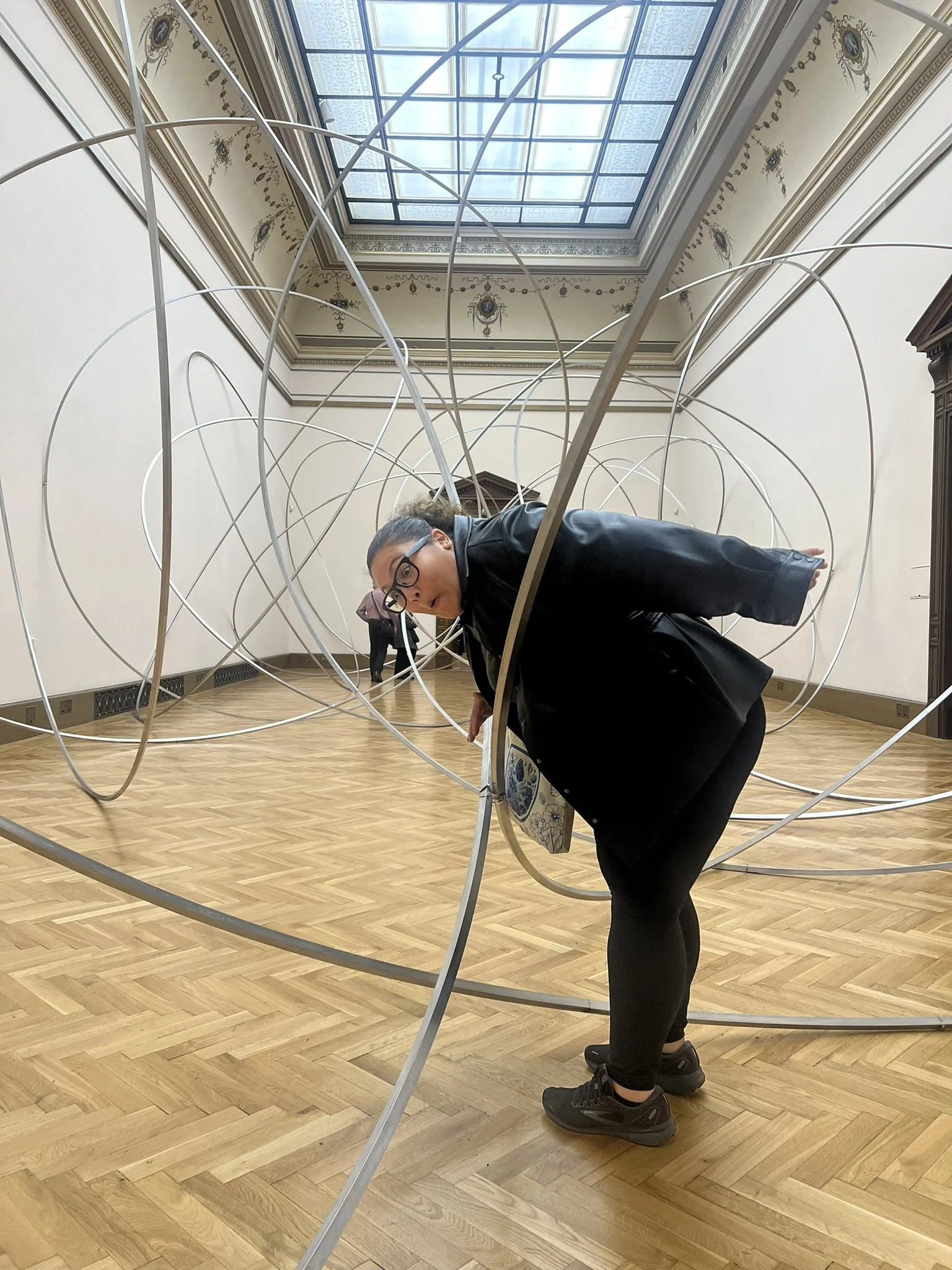Person with glasses leaning through an art installation of metal loops inside a gallery with a wooden floor and a glass ceiling.